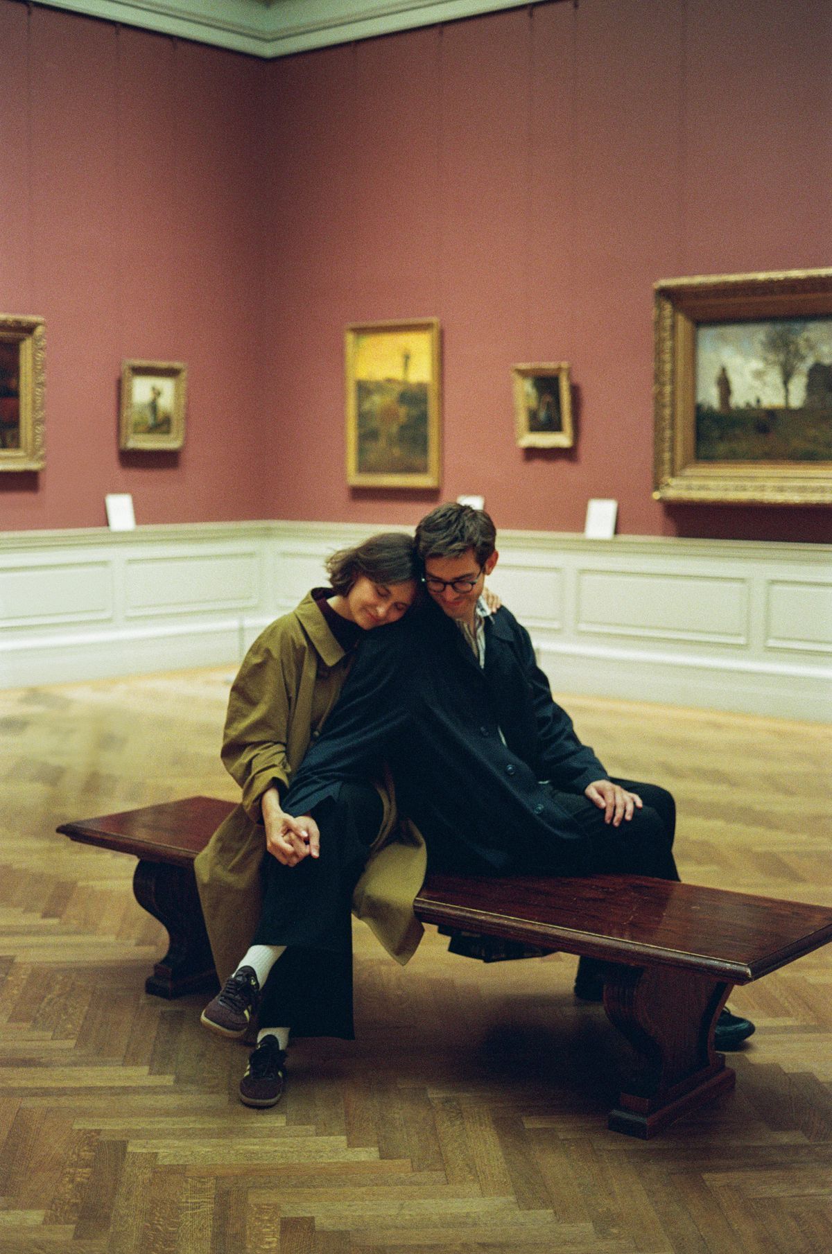Couple sitting closely on a wooden bench in a museum gallery with pink walls and landscape paintings.