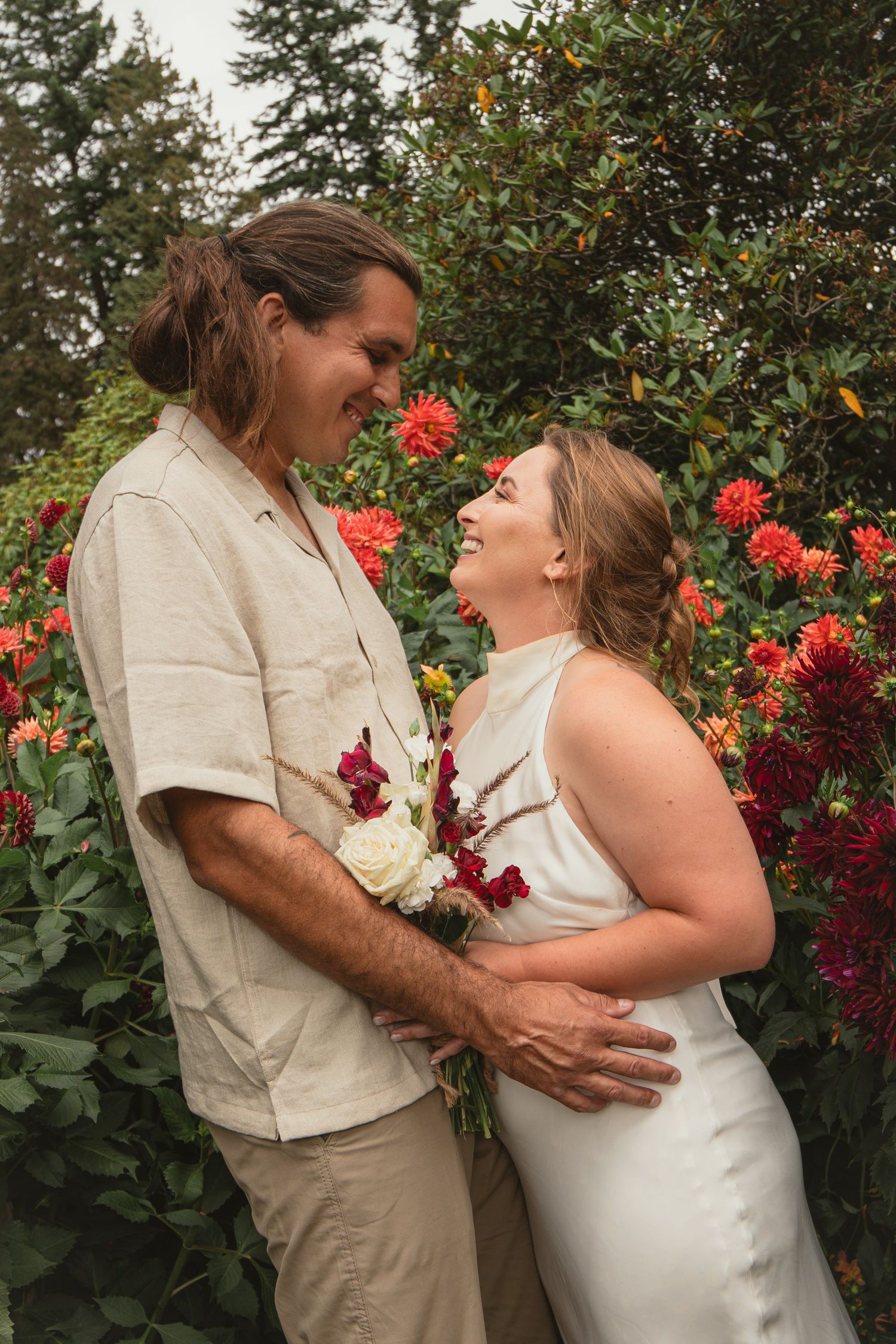 A couple smiles at each other holding a bouquet in front of vibrant red dahlias, dressed in wedding attire.