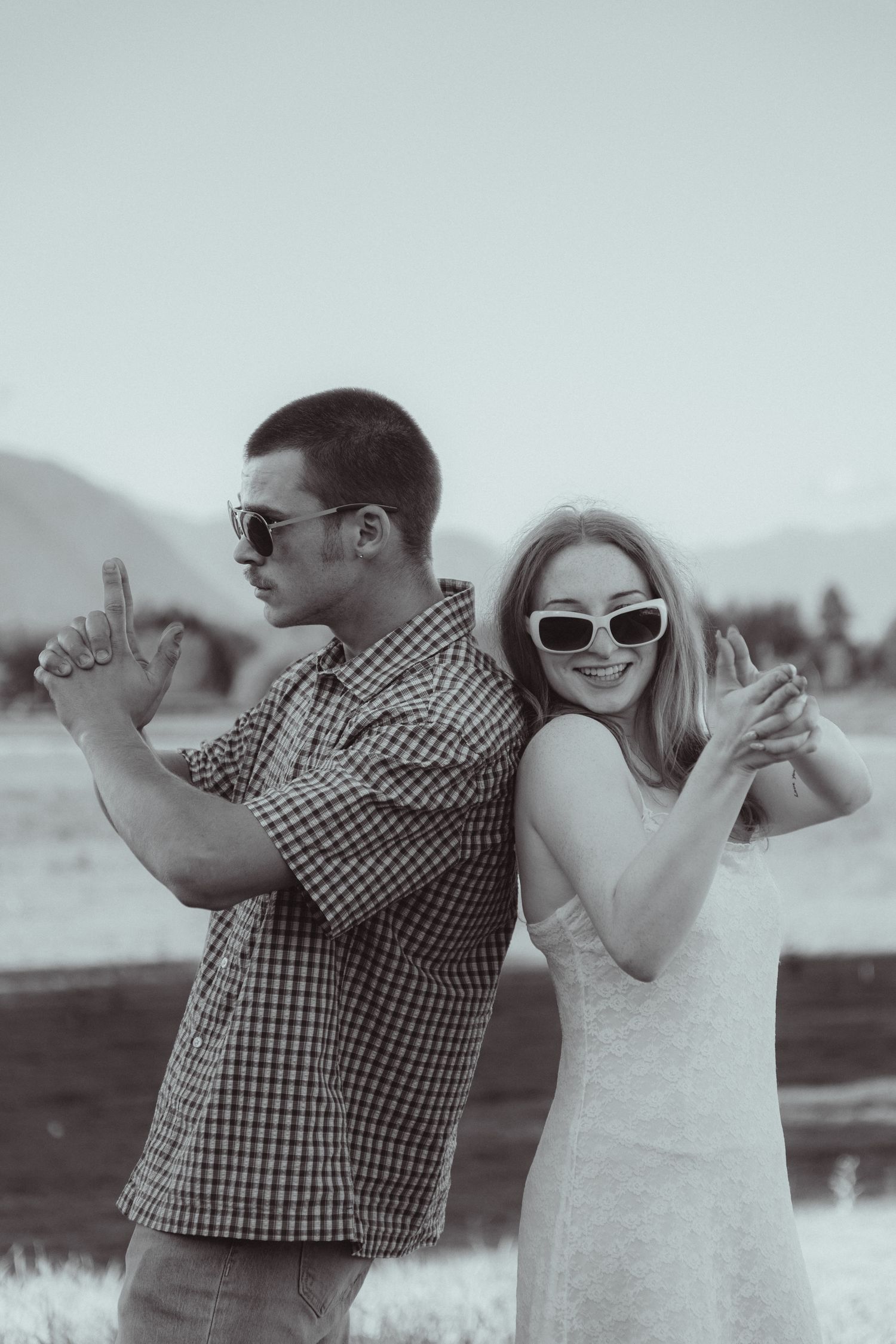 A couple in sunglasses pose back-to-back outdoors, smiling and making fun hand gestures in a black and white photo.