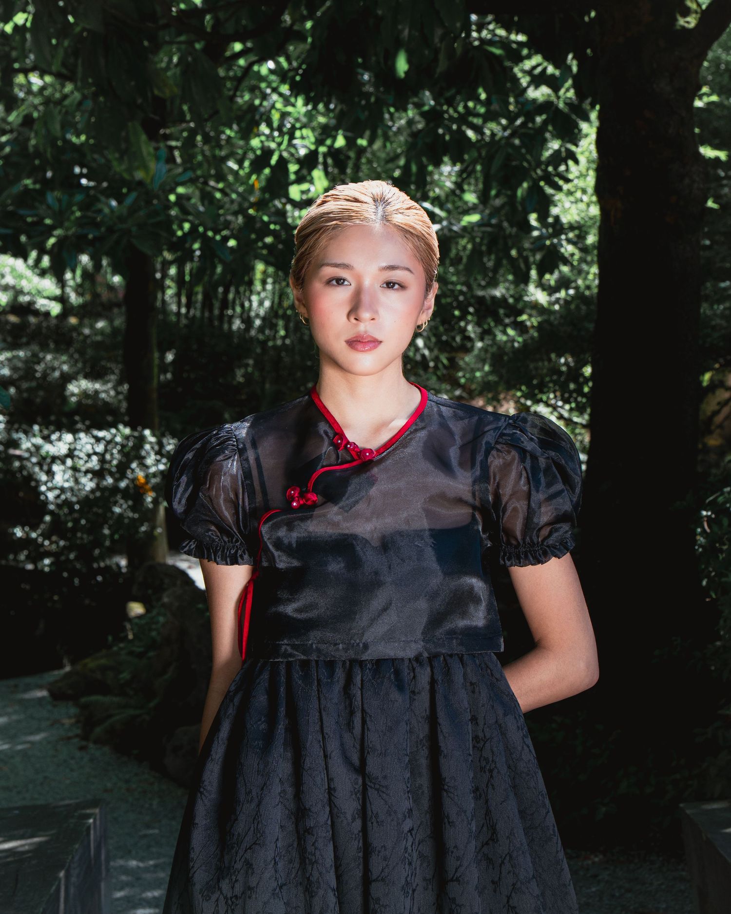 Young woman in black lace dress with red trim stands in lush green garden setting.