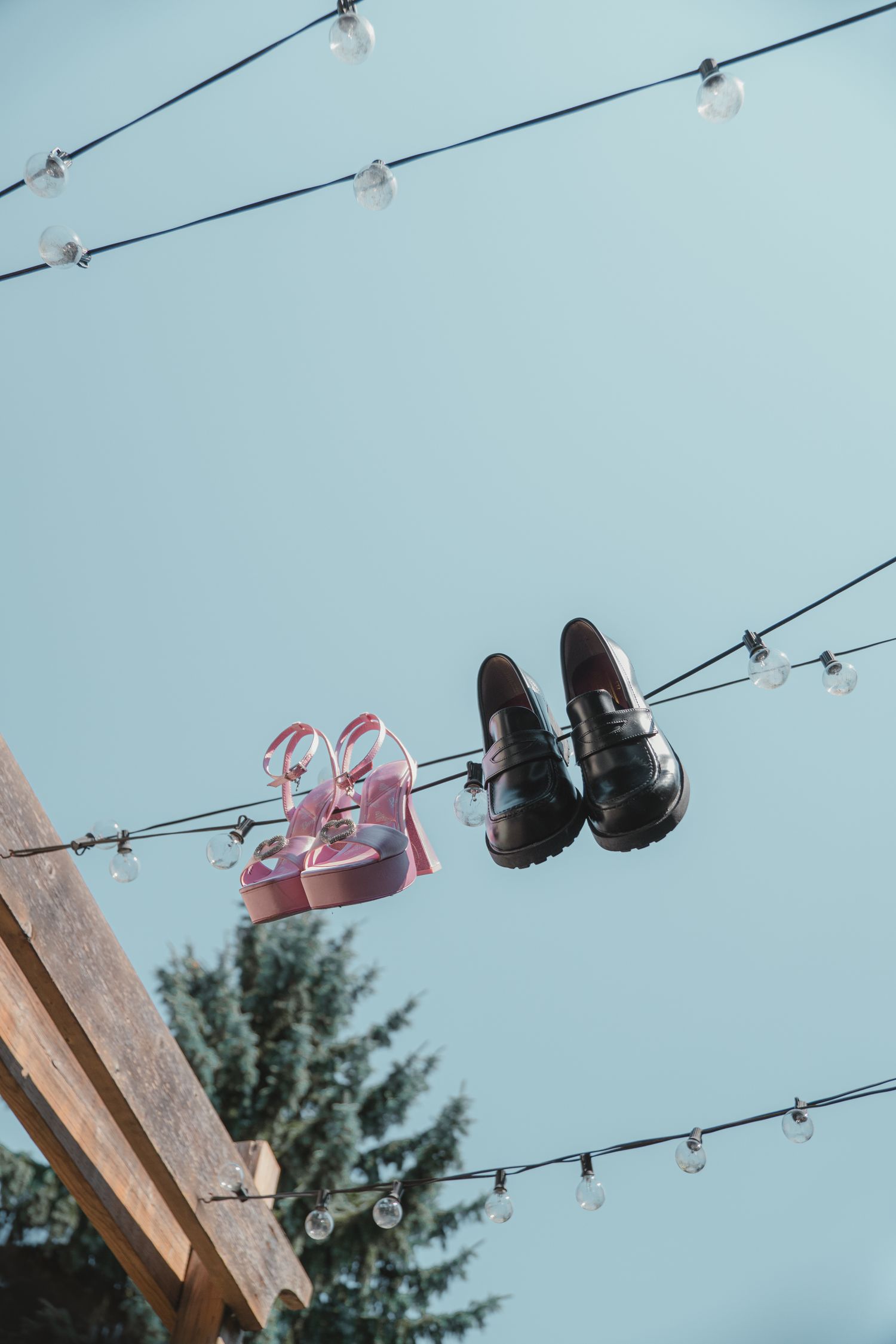 Pink heels and black loafers hanging on string lights against a clear blue sky near a wooden beam.