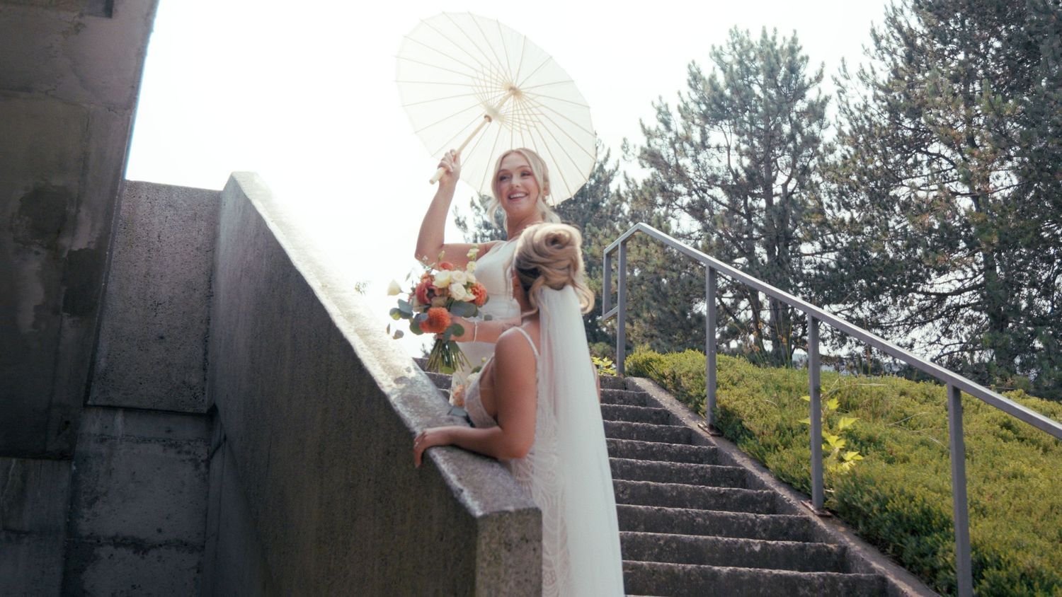 Elegant bride in white gown holding parasol and bouquet on stone steps outdoors.