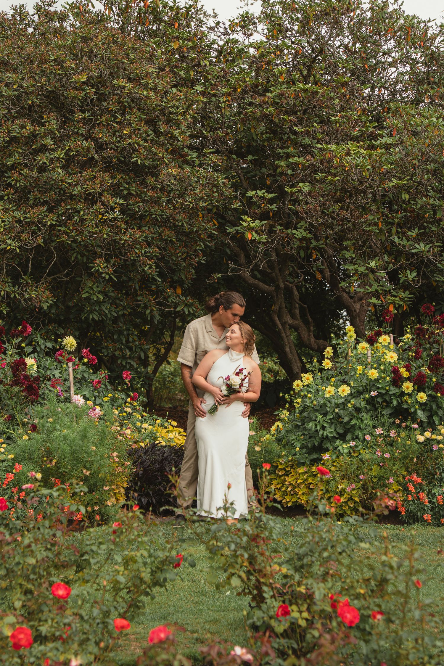 Bride and groom embrace among vibrant rose garden blooms, surrounded by lush greenery in an outdoor wedding portrait.