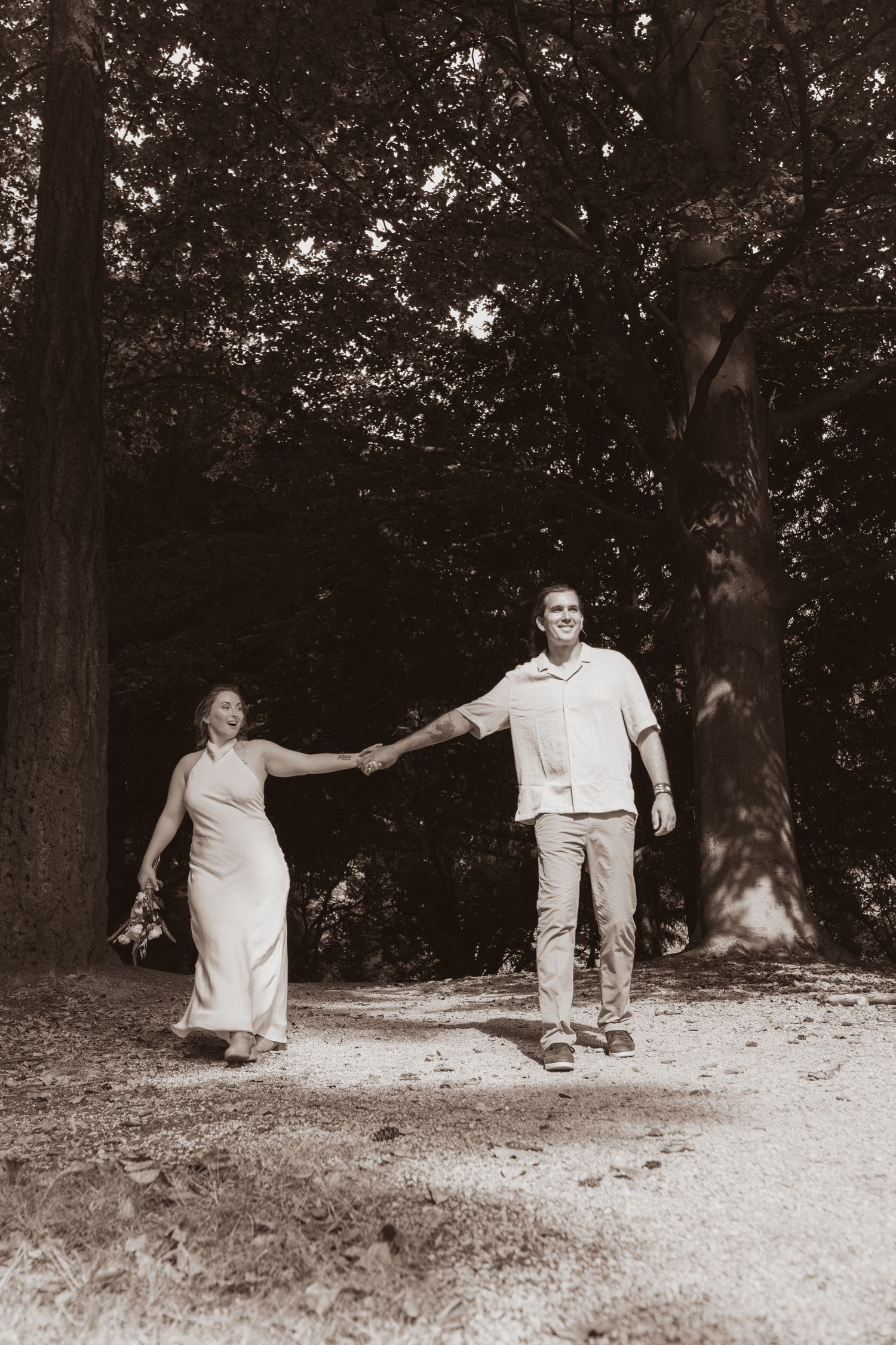 Sepia-toned photo of a smiling couple holding hands while walking on a forest trail.