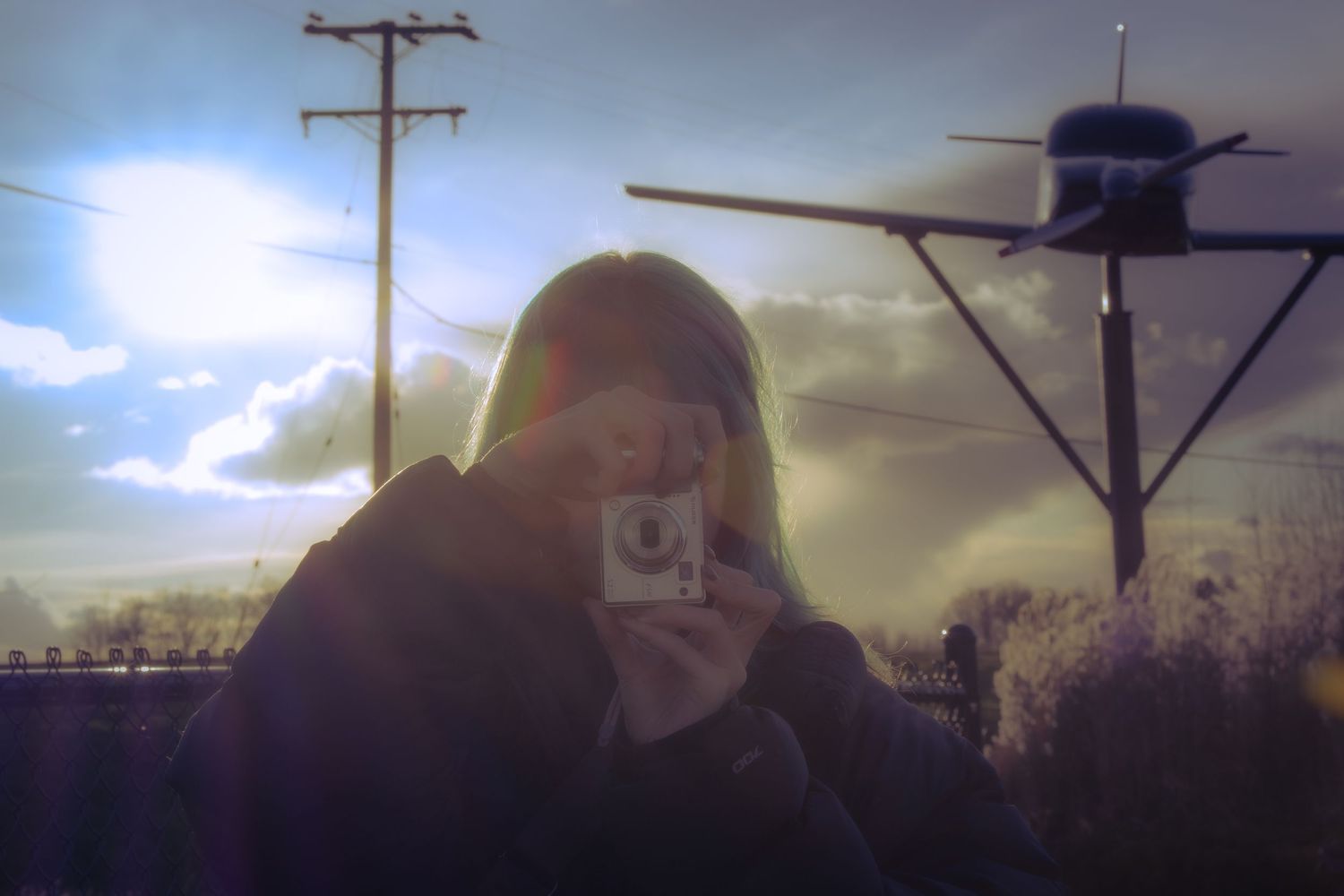 Person photographing with instant camera outdoors with dramatic cloudy sky, power lines, and lens flare effect.
