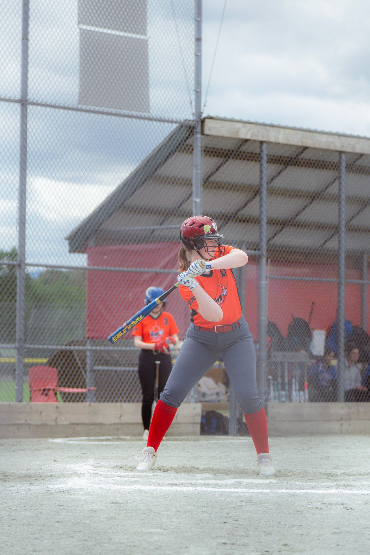 Young female softball batter in orange and red uniform stands ready at home plate.
