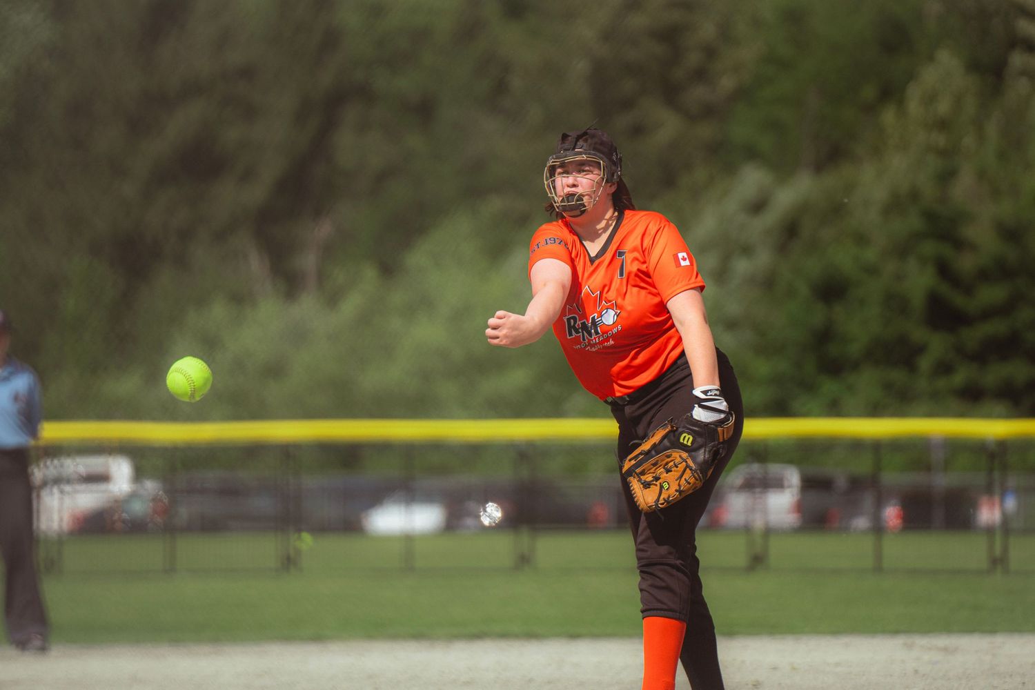 Female softball pitcher in orange uniform releasing a yellow ball during a game.