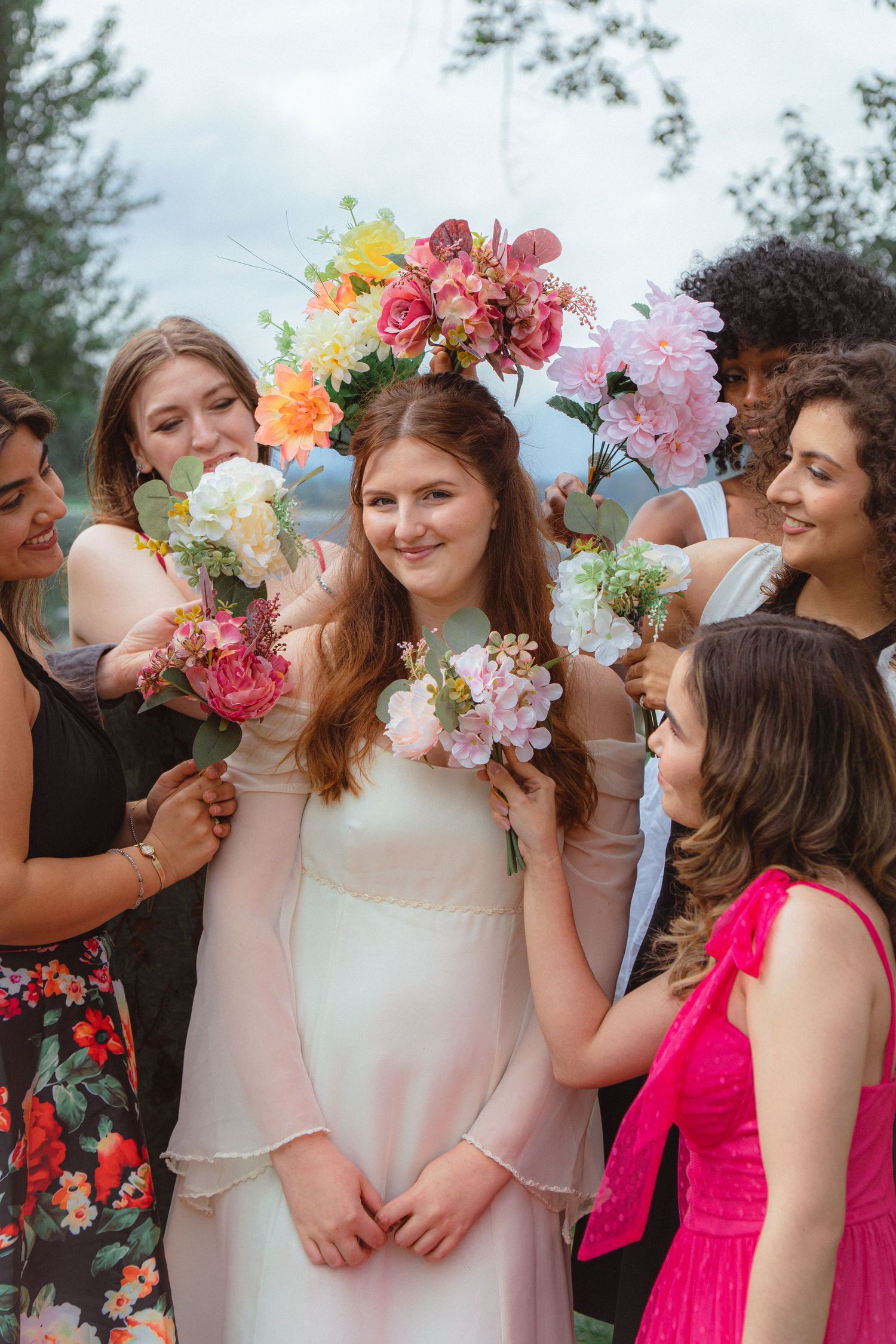 Bride in white dress surrounded by bridesmaids holding colorful flower bouquets at outdoor wedding.