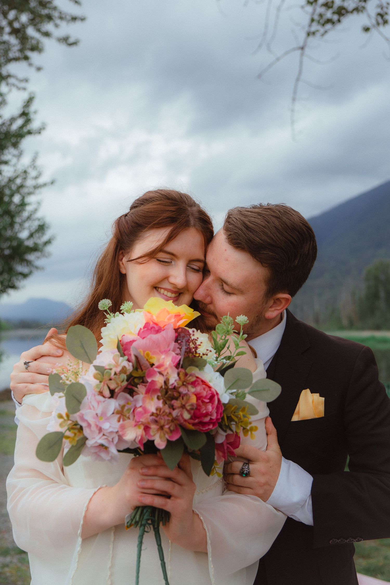 Happy bride laughing as groom kisses her cheek while holding vibrant floral bouquet by lakeside.