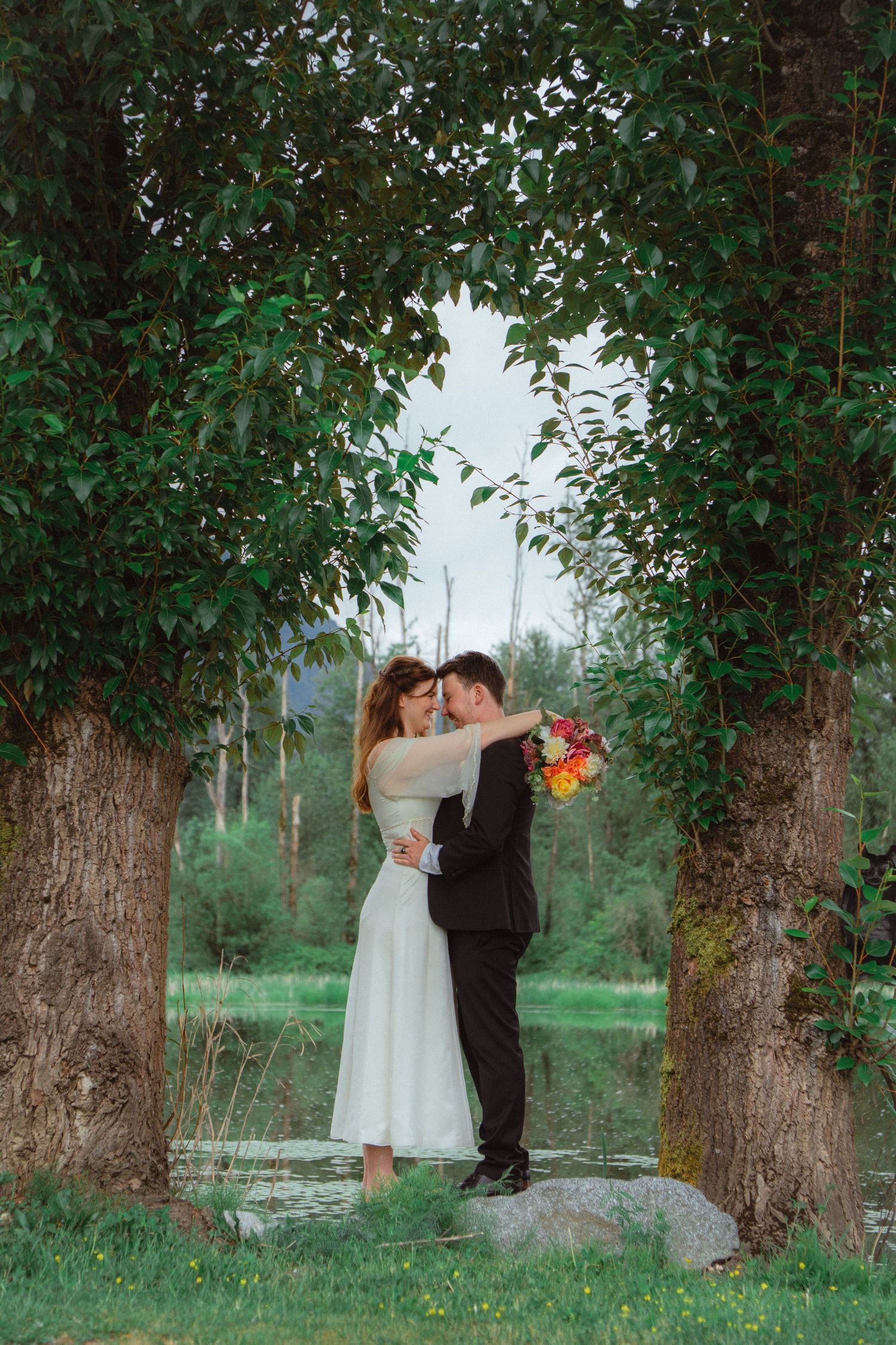 Bride and groom embrace romantically under a lush green tree arch in a scenic outdoor setting.