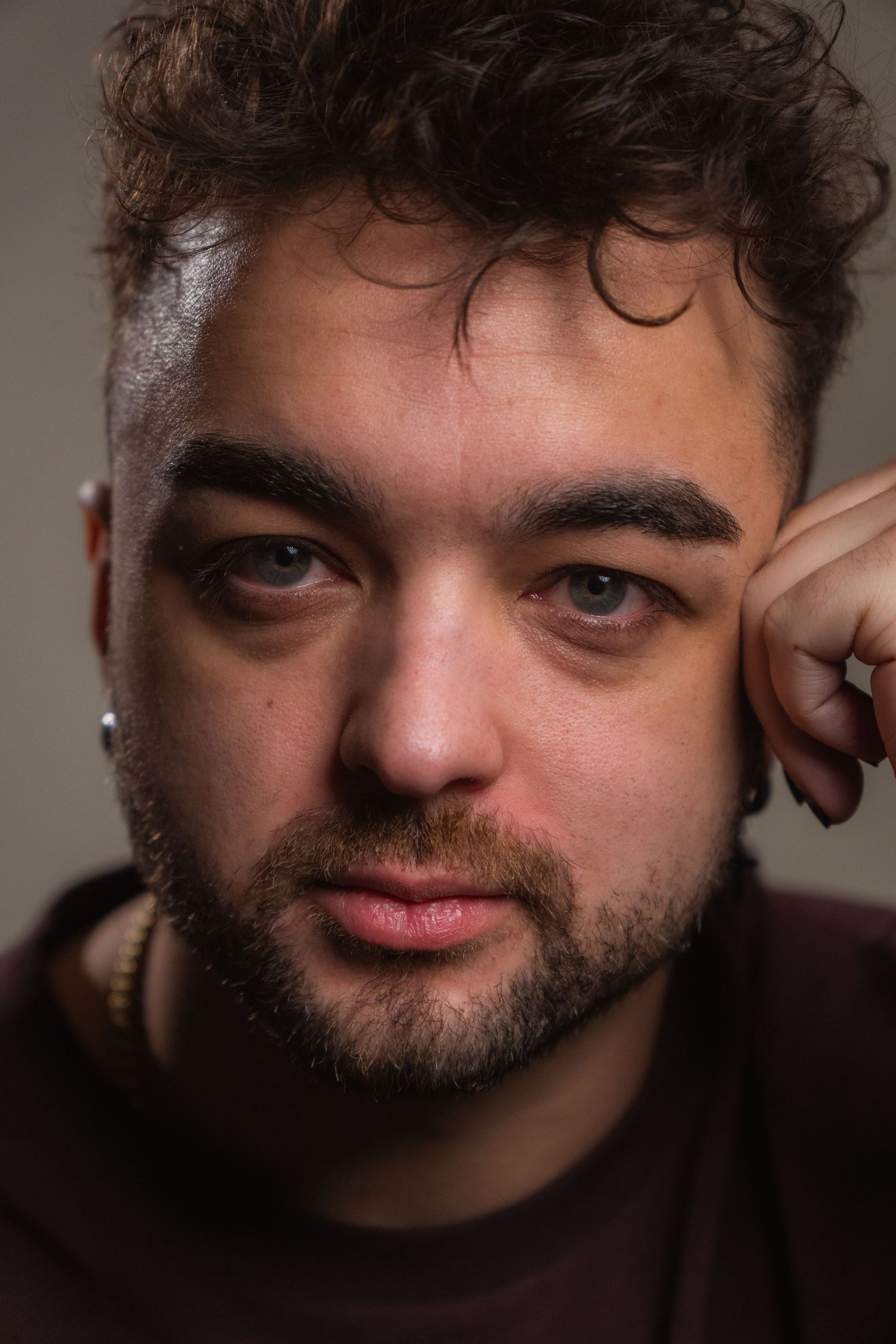 Close-up portrait of bearded young man with curly hair resting hand near face, intense gaze, studio lighting.