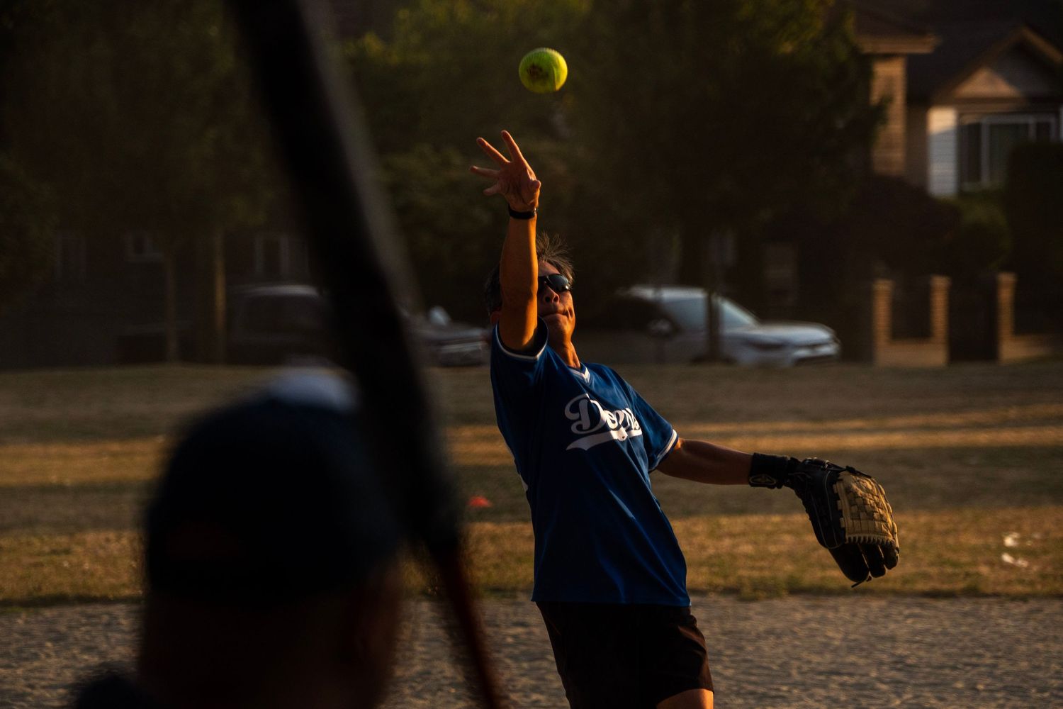 Softball pitcher in blue uniform releases a yellow ball during a game in warm evening sunlight.