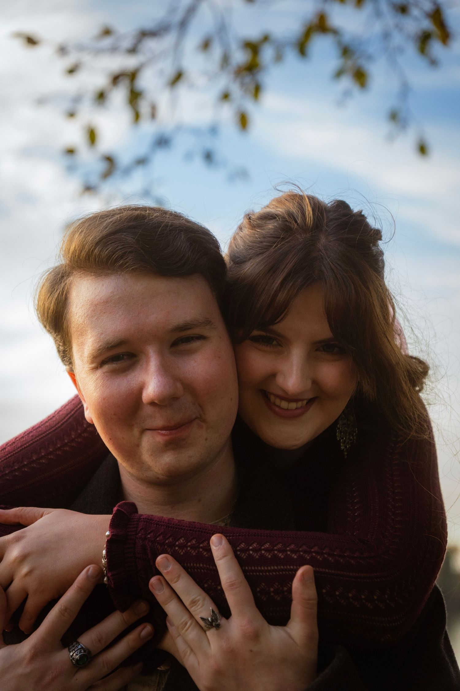 Happy young couple posing together outdoors, woman hugging man from behind, cloudy sky background.