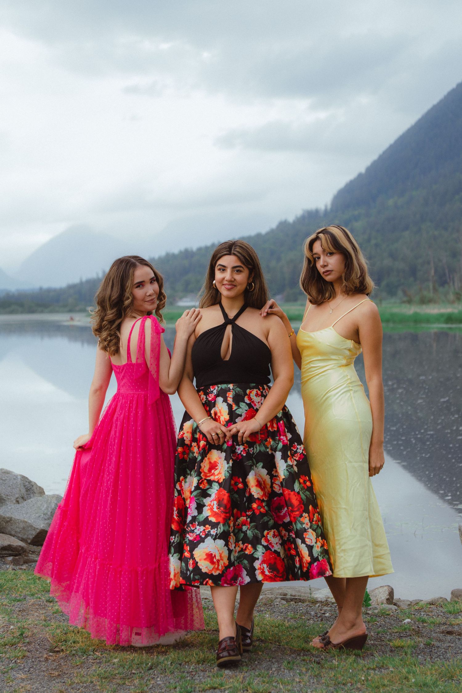 Three women in elegant dresses pose by a mountain lake on a cloudy day.