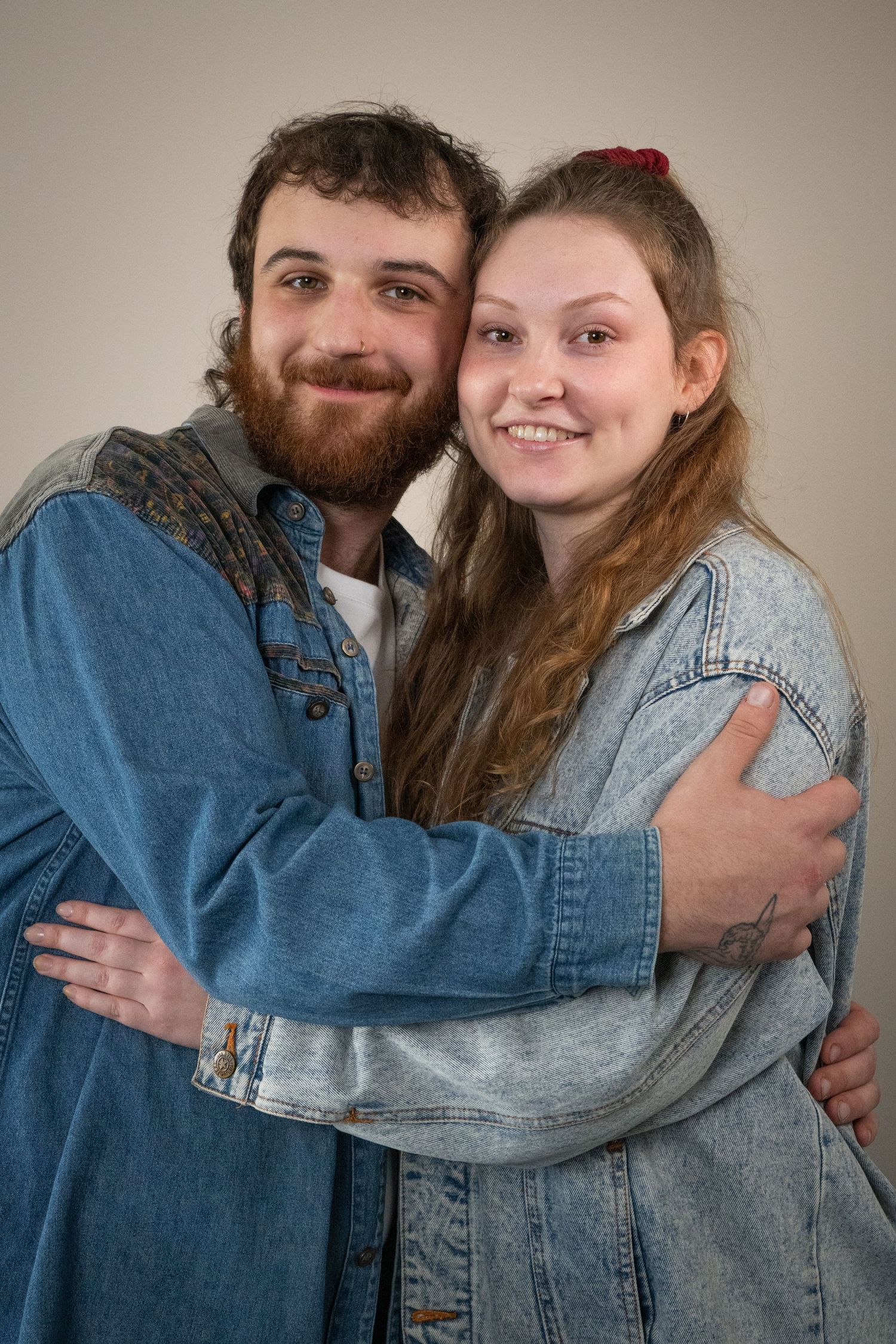 A smiling couple wearing coordinating denim jackets hug each other in a casual portrait against a neutral wall.