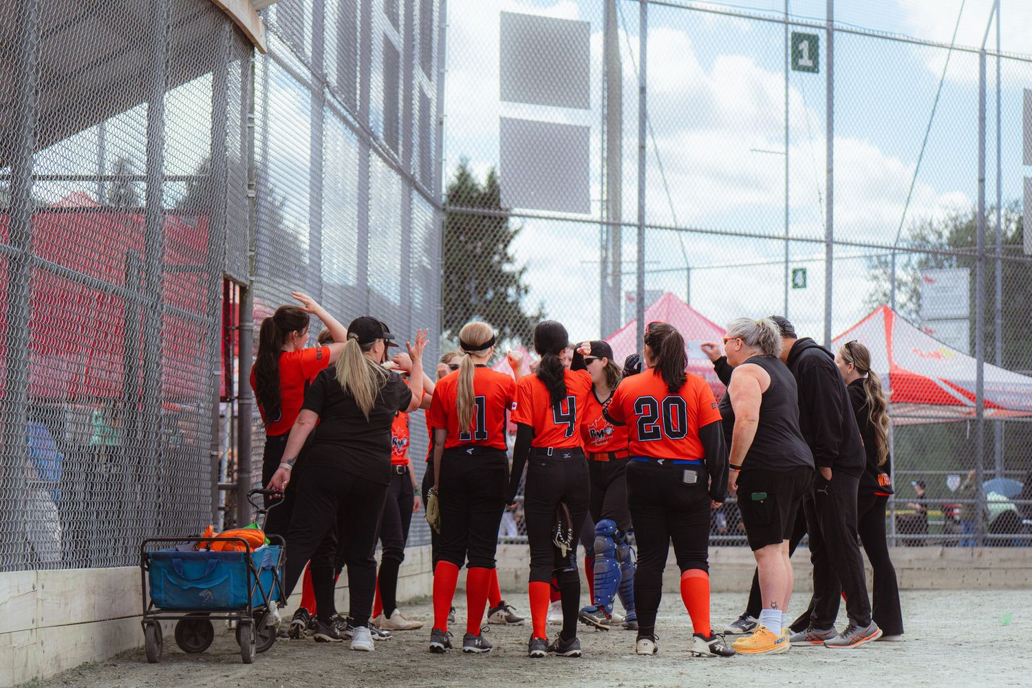 Women's softball team in orange jerseys huddle near a chain-link fence during an outdoor tournament.