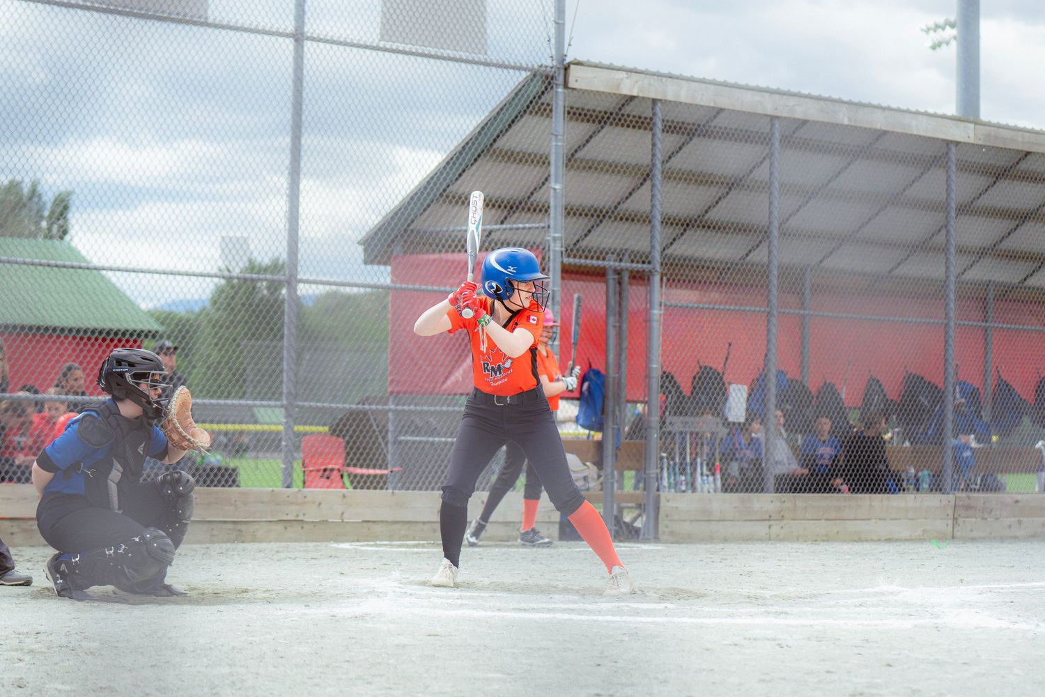Female softball player in orange uniform bats while catcher and crowd watch from behind.