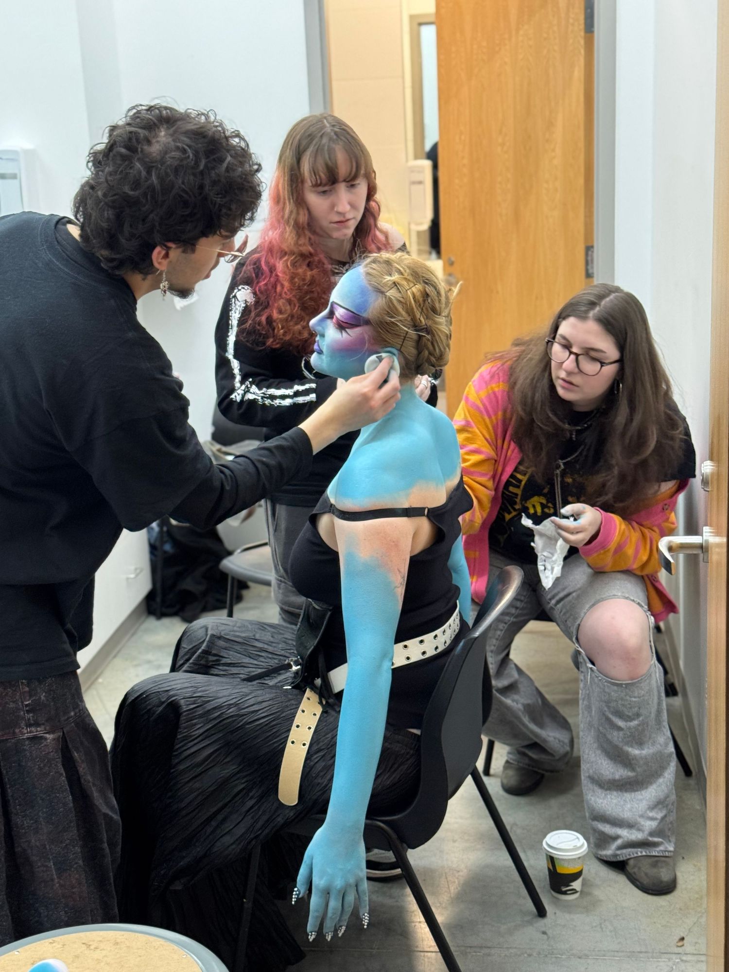 Three makeup artists apply elaborate blue body paint to a model in a backstage preparation area.