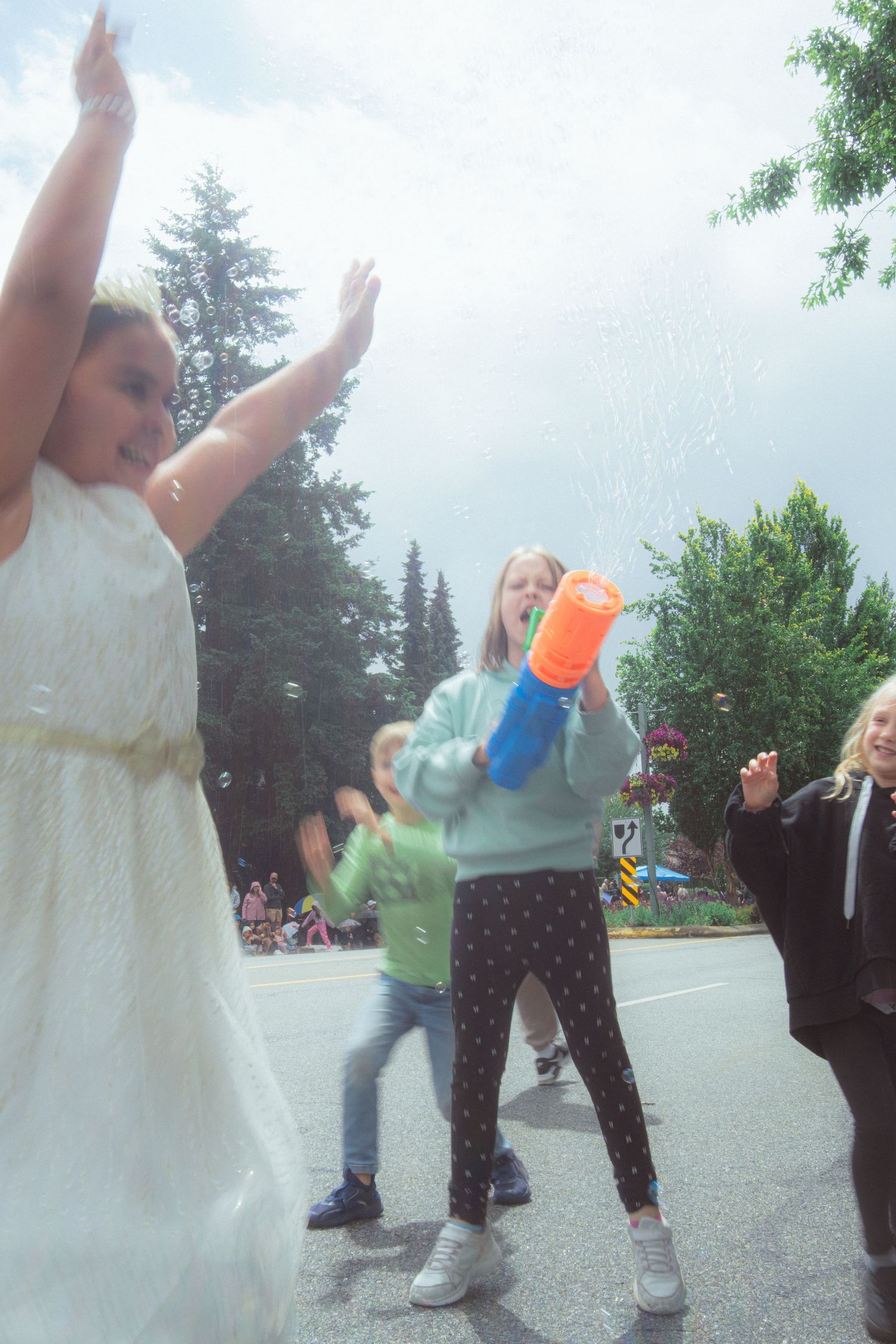 Children play with a bubble gun during a street parade on an overcast day, with trees in the background.