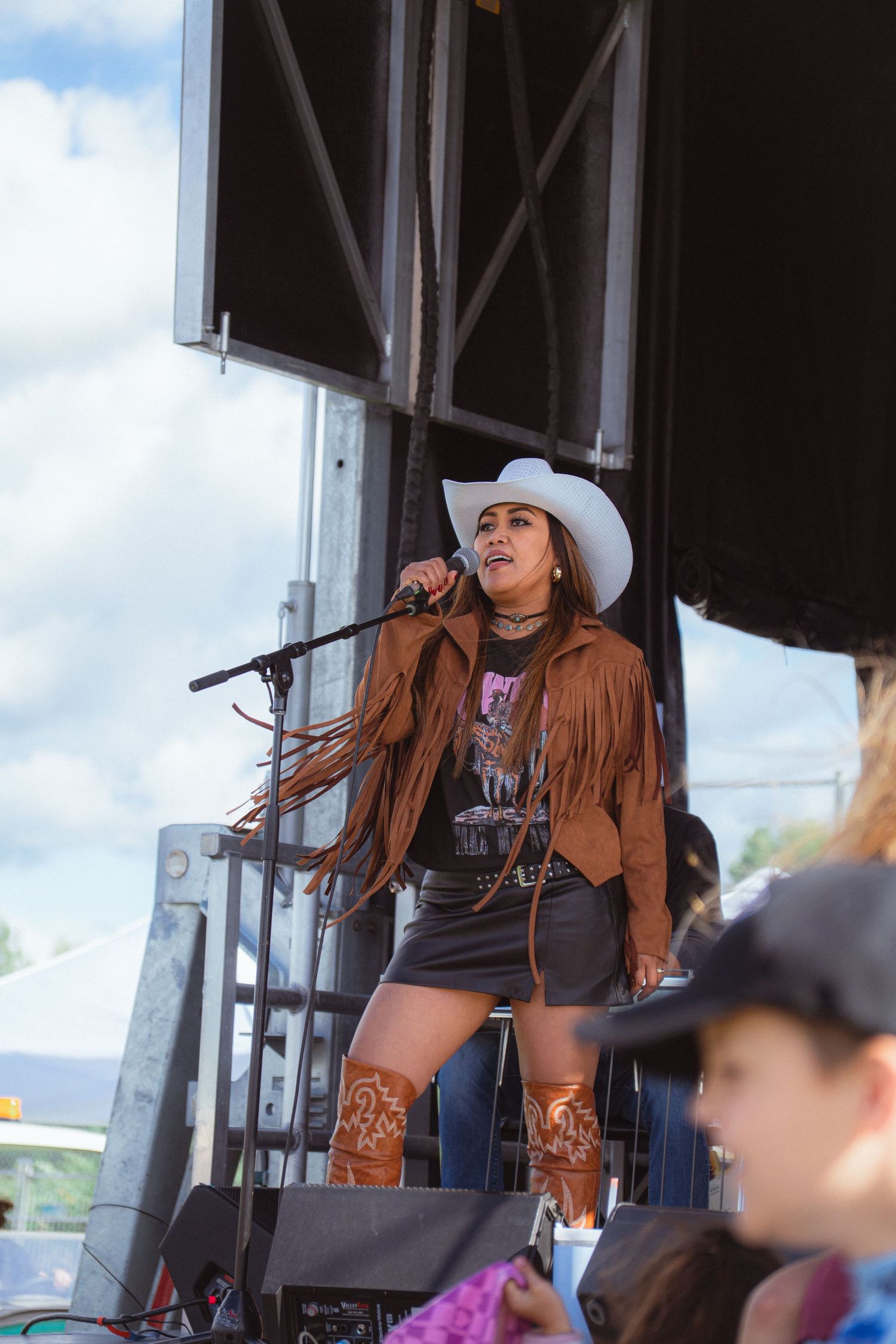 Female country singer performing on stage in white cowboy hat and fringed brown jacket with mic.