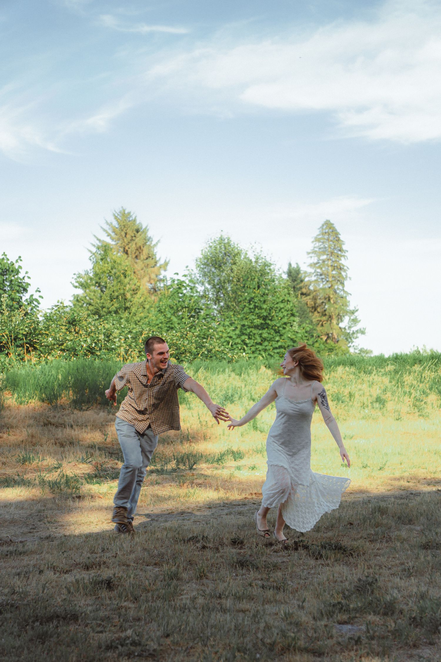 A joyful couple runs and dances together in a sunny green field, the woman in a white dress and man in casual attire.