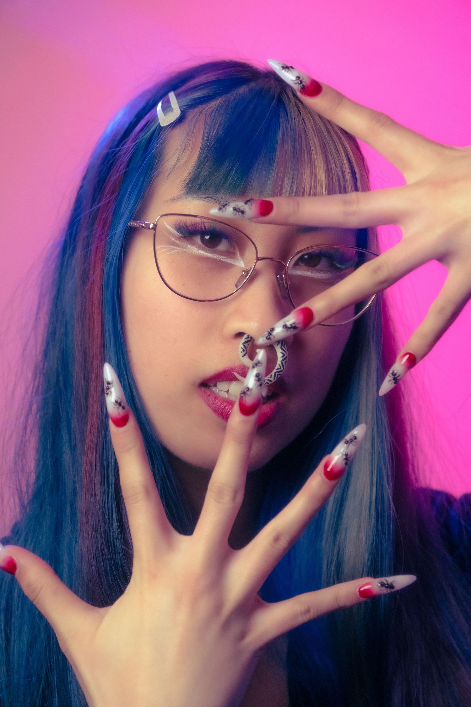 Young Asian woman with blue hair, glasses, and long decorated nails poses against a pink background.