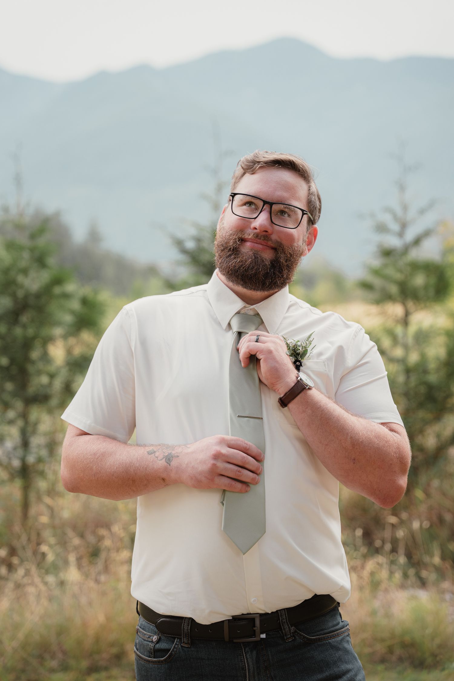 Bearded groom in white shirt and sage tie adjusting boutonniere with mountain backdrop.