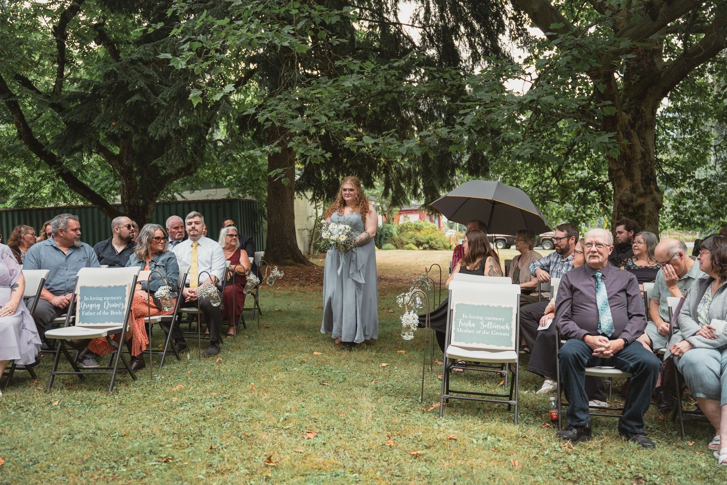 Outdoor wedding ceremony on green lawn with guests seated in chairs, bridesmaid walking aisle, large trees providing shade.