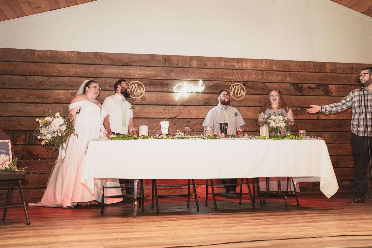 Wedding party stands and sits at a decorated head table with floral arrangements in a rustic barn reception venue.