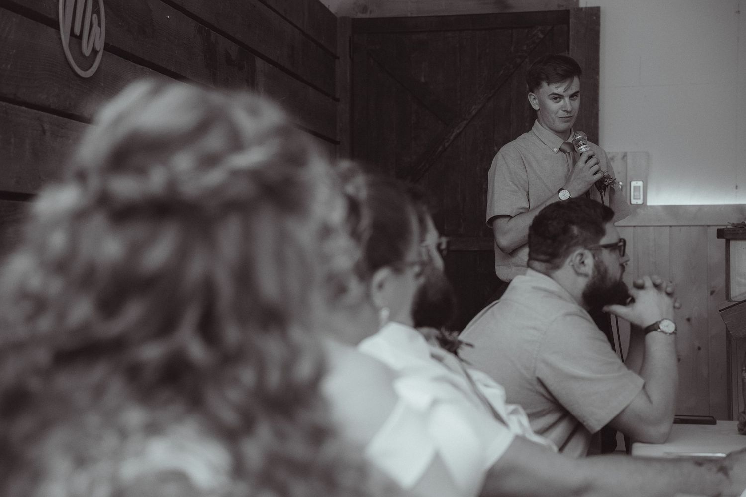 Black and white photo of man giving speech at wedding reception with guests seated at tables in rustic venue.