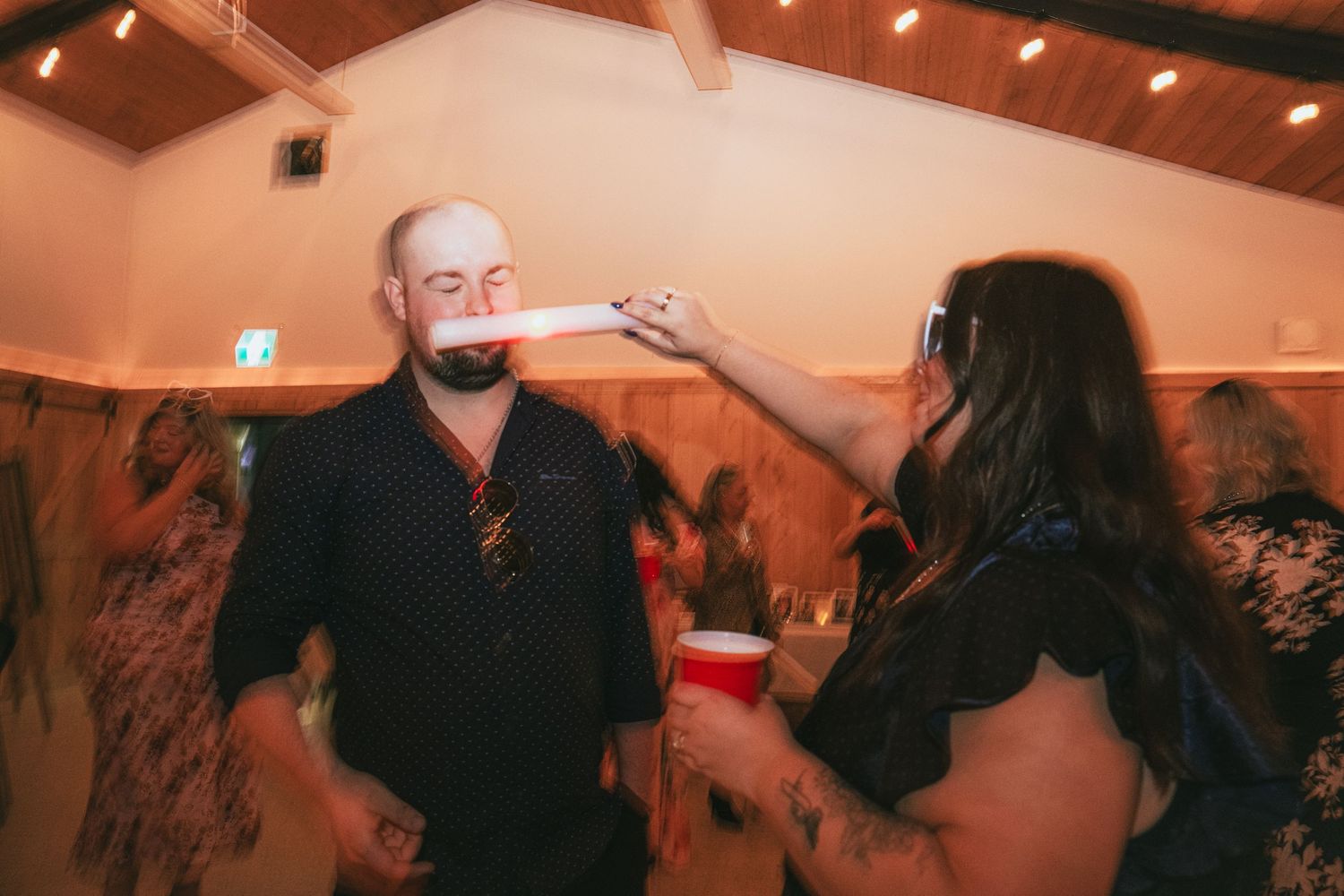 Two people at an indoor party event, one holding a red cup near the other's face in a dimly lit venue.