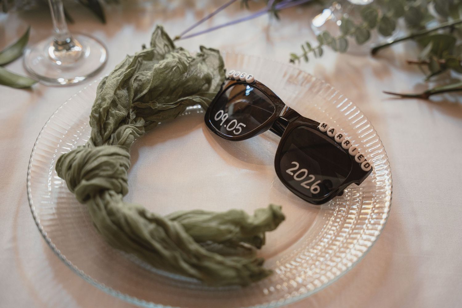 Sunglasses engraved with 2026 date on a glass plate with sage green silk ribbon wedding table setting.