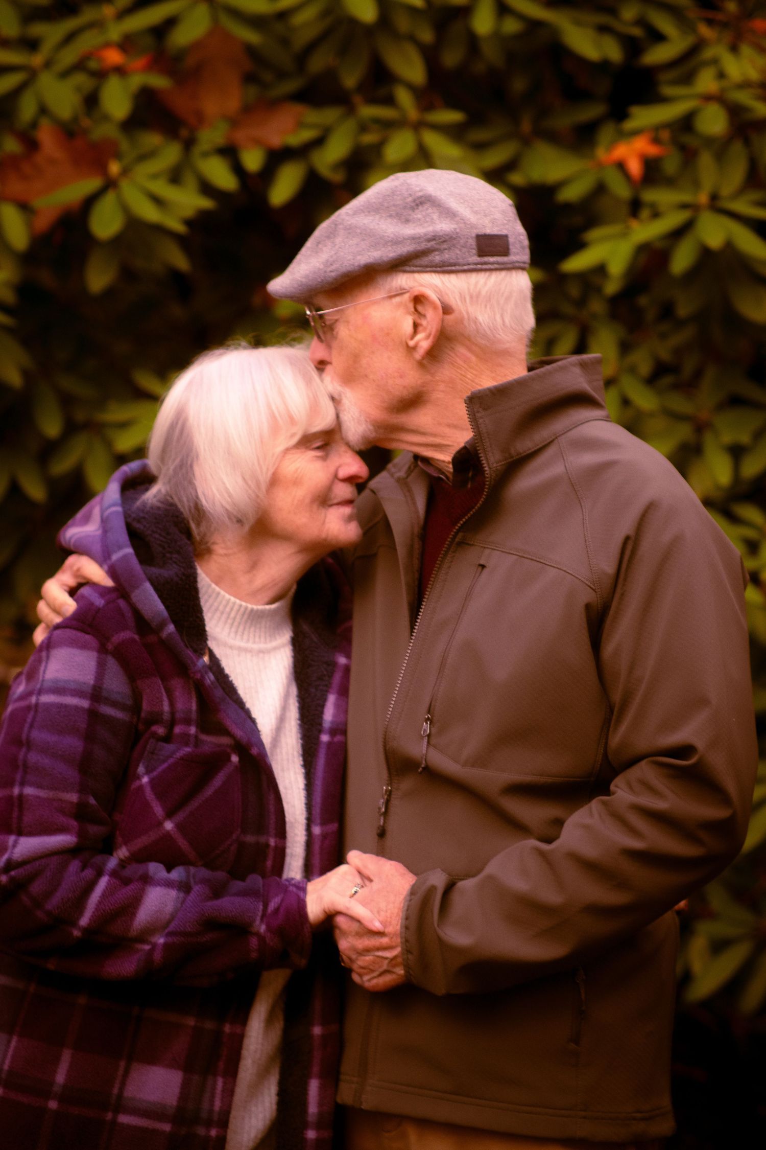 Elderly man tenderly kissing woman on forehead while holding hands in front of lush green foliage.