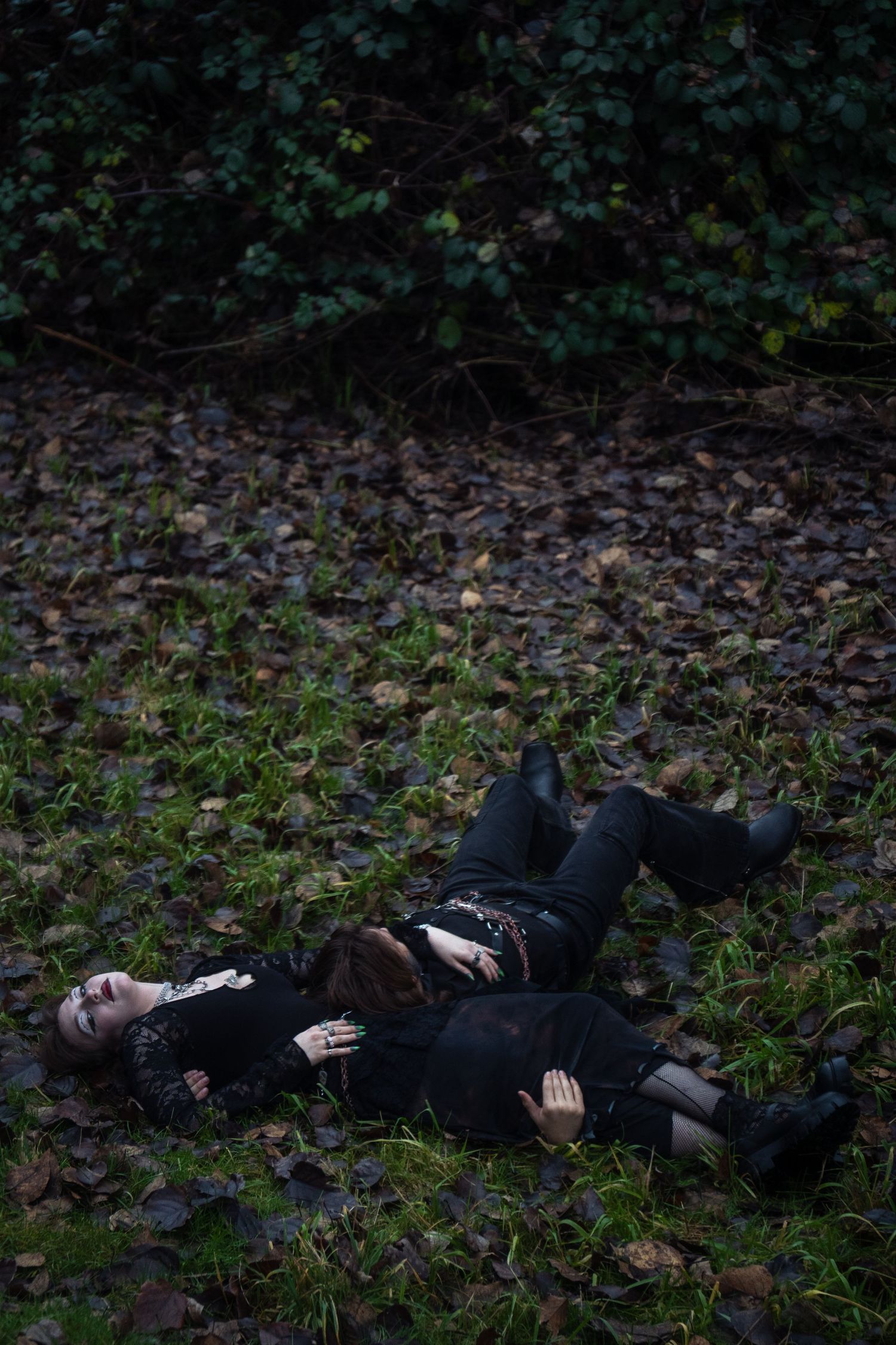 Two people dressed in black lie together on mossy ground near dark foliage.