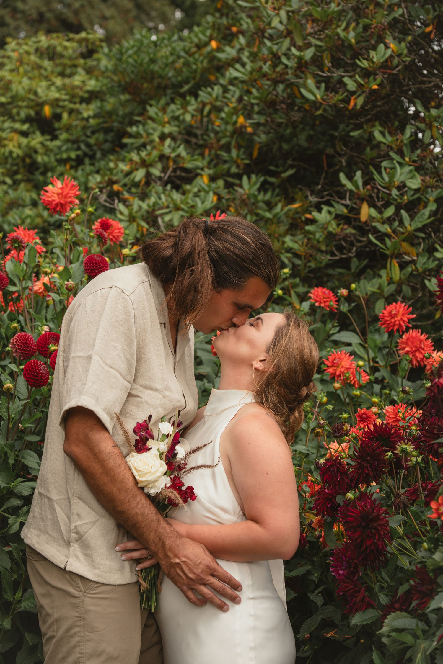 Bride and groom sharing a kiss among lush red dahlias in an outdoor garden setting.