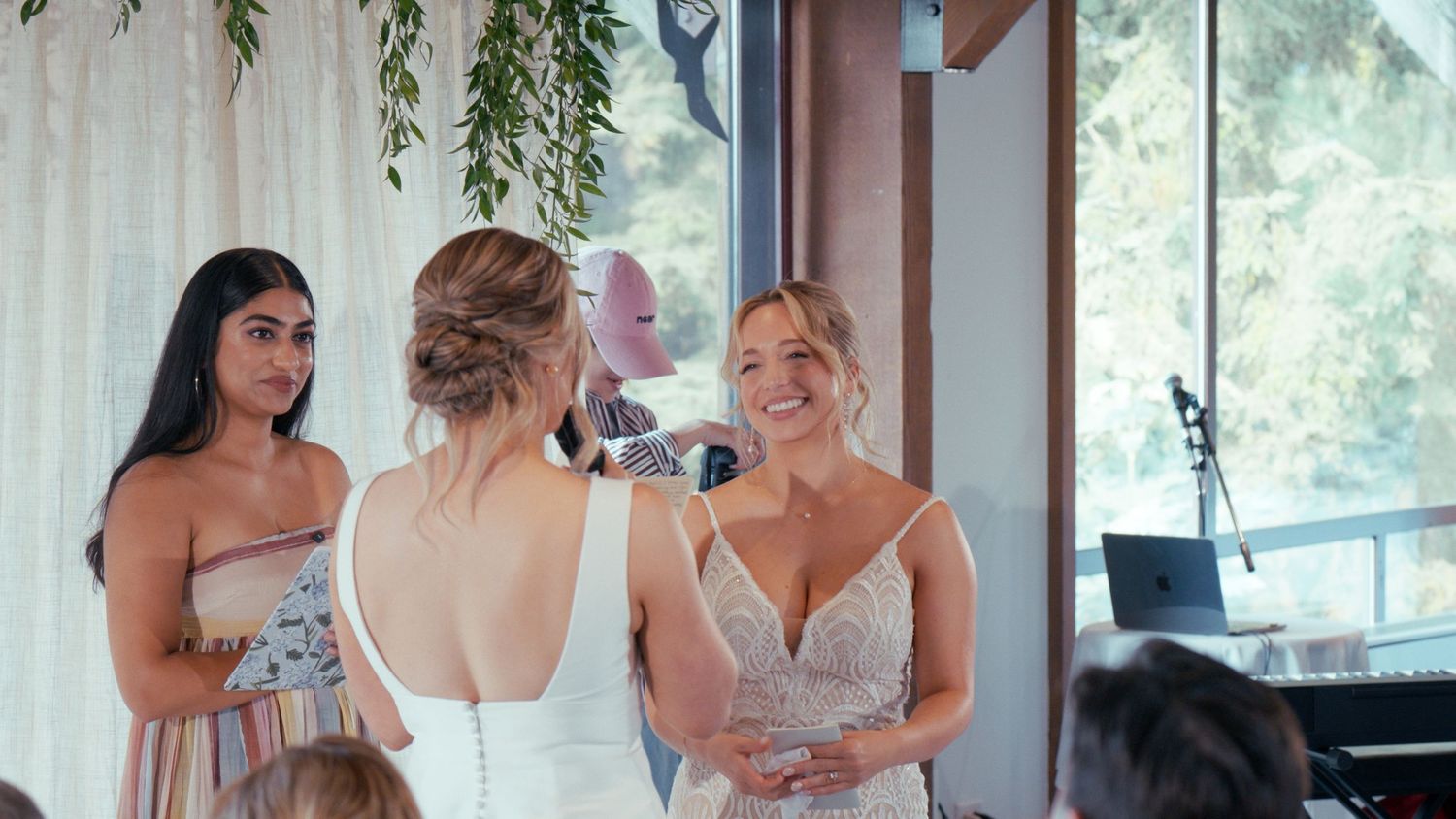 Bride in white lace gown smiles with bridesmaids in floral dresses at elegant indoor wedding venue.