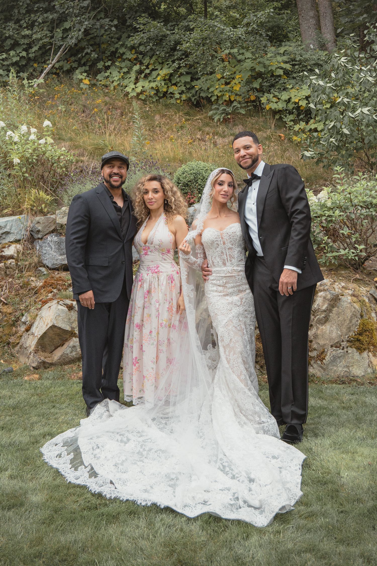 Two couples pose outdoors at a wedding, bride in lace gown with veil, groom in tuxedo, guests in formal attire.
