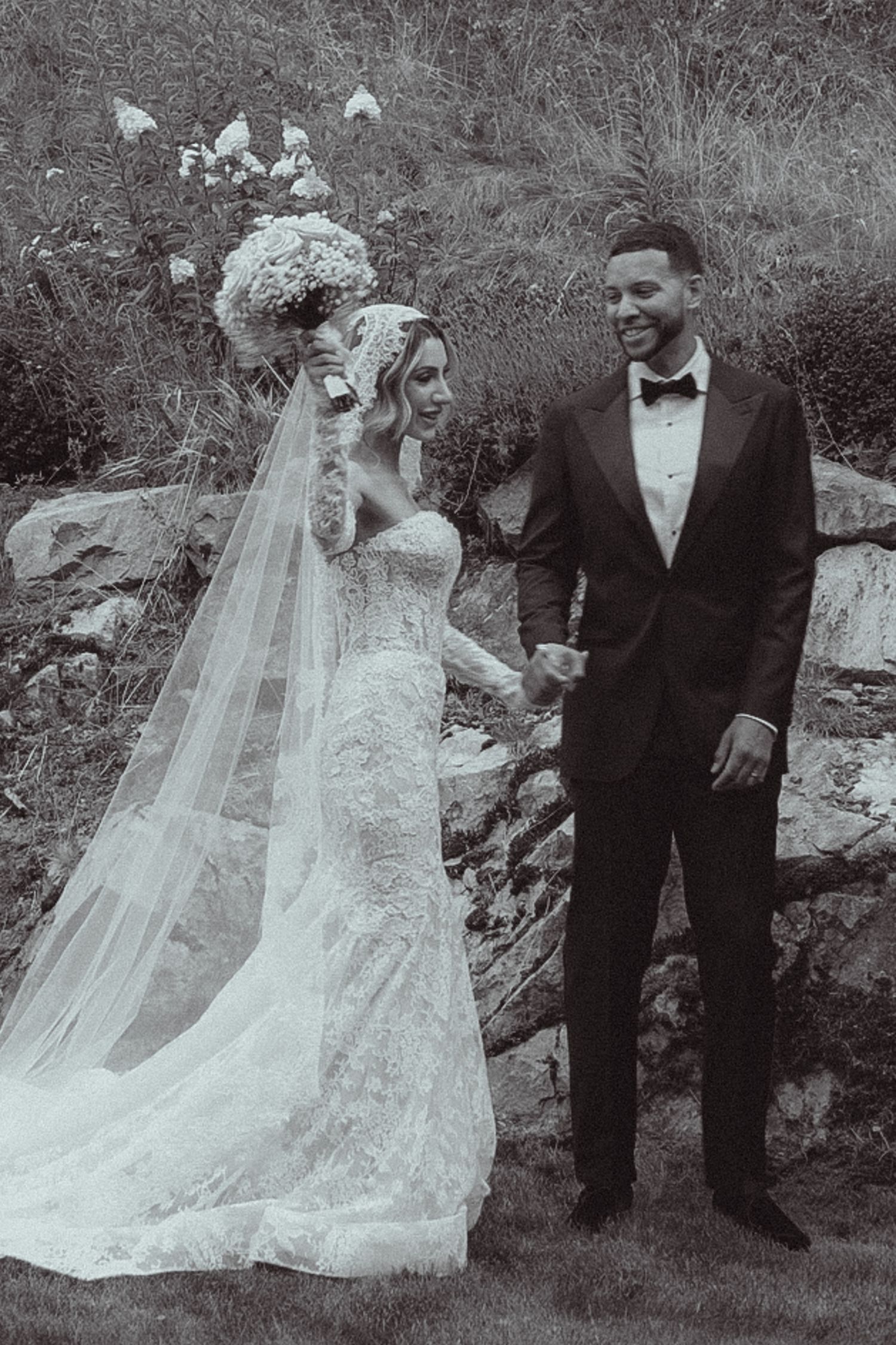 Black and white photo of a bride in lace gown holding bouquet and groom in tuxedo holding hands at outdoor wedding.