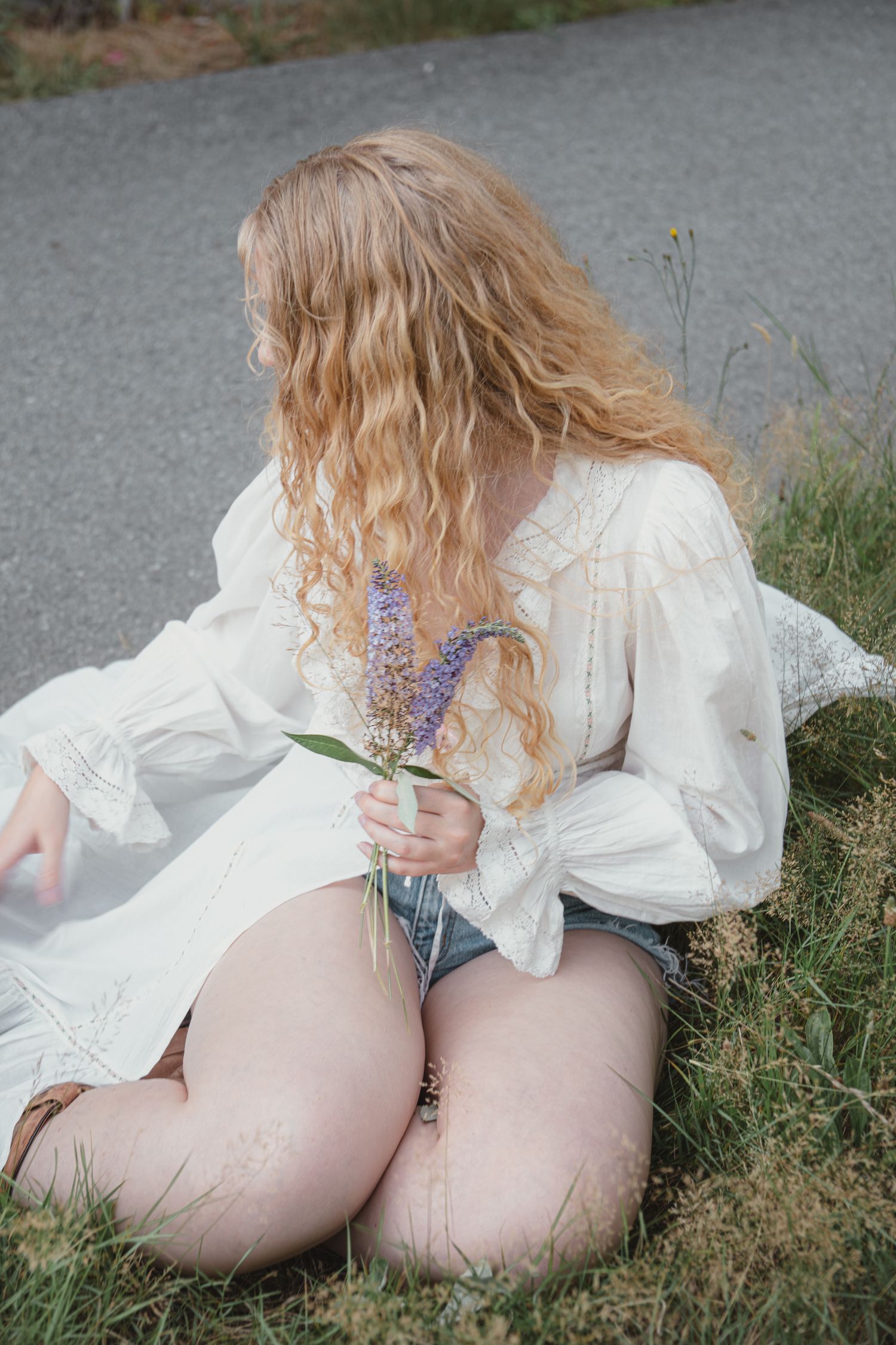 Curly blonde woman in white blouse and denim shorts sitting on grass holding purple wildflowers near road.