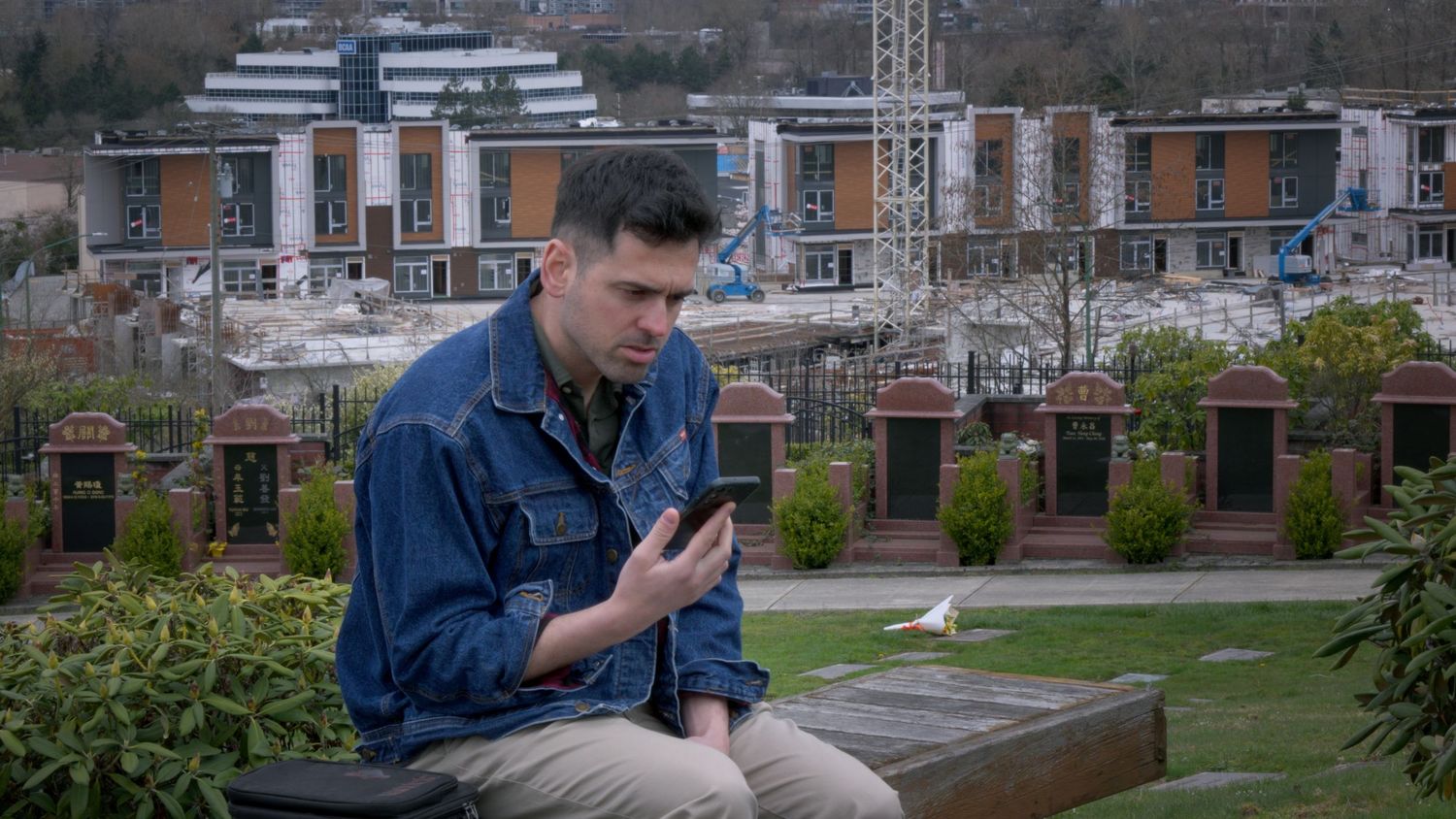 Young man in denim jacket sitting in a cemetery using a smartphone with city buildings in background.