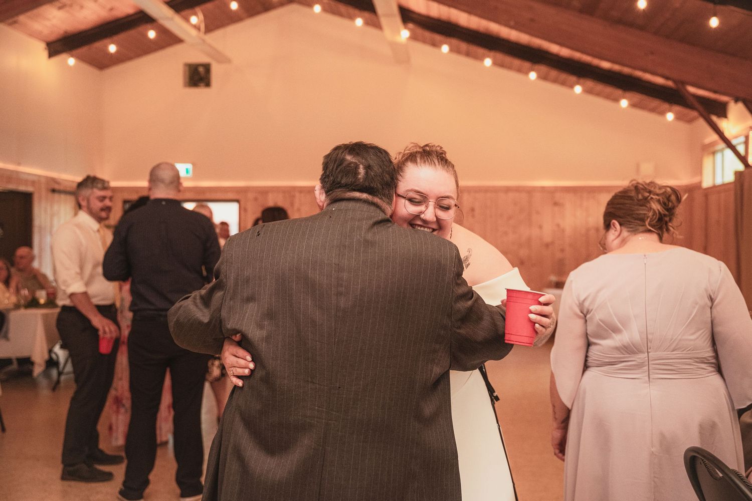 Bride hugging a guest in a suit at a wedding reception in a warmly lit barn venue.