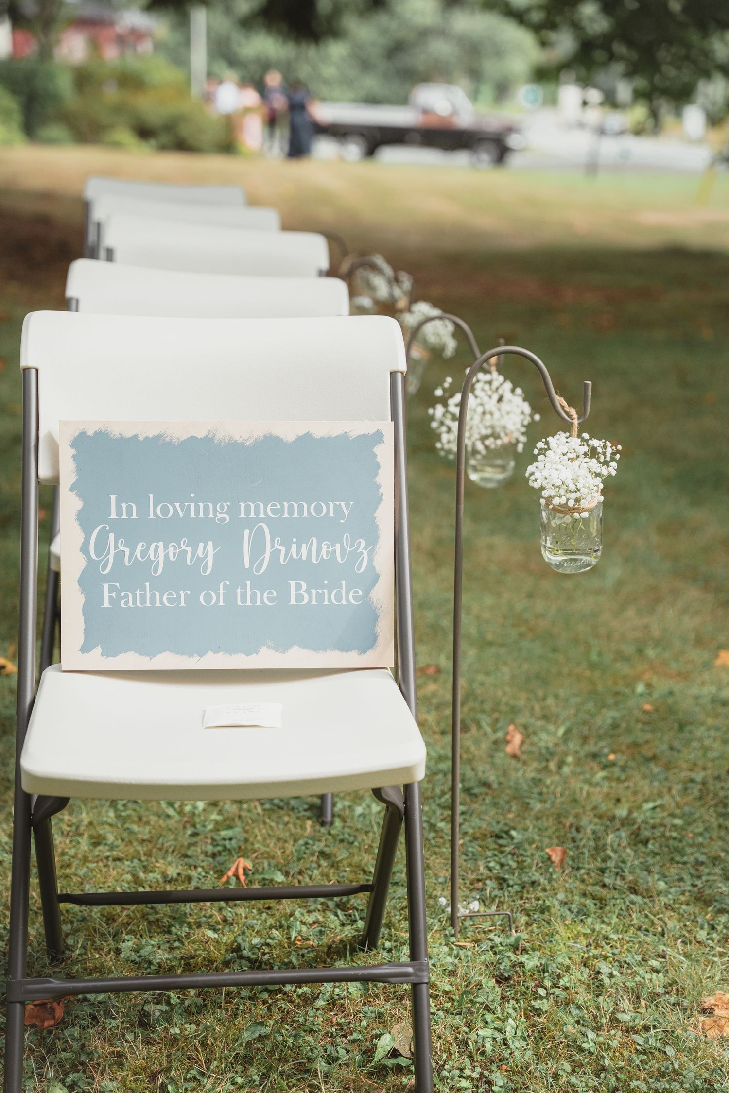 White folding chair with blue memorial sign for Gregory Drinovic, Father of the Bride, beside baby's breath flowers at outdoor wedding.