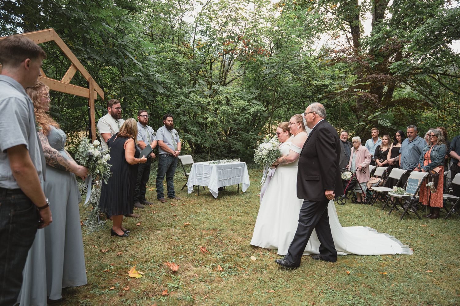 Bride walks down outdoor aisle with escort at rustic woodland wedding ceremony with seated guests.