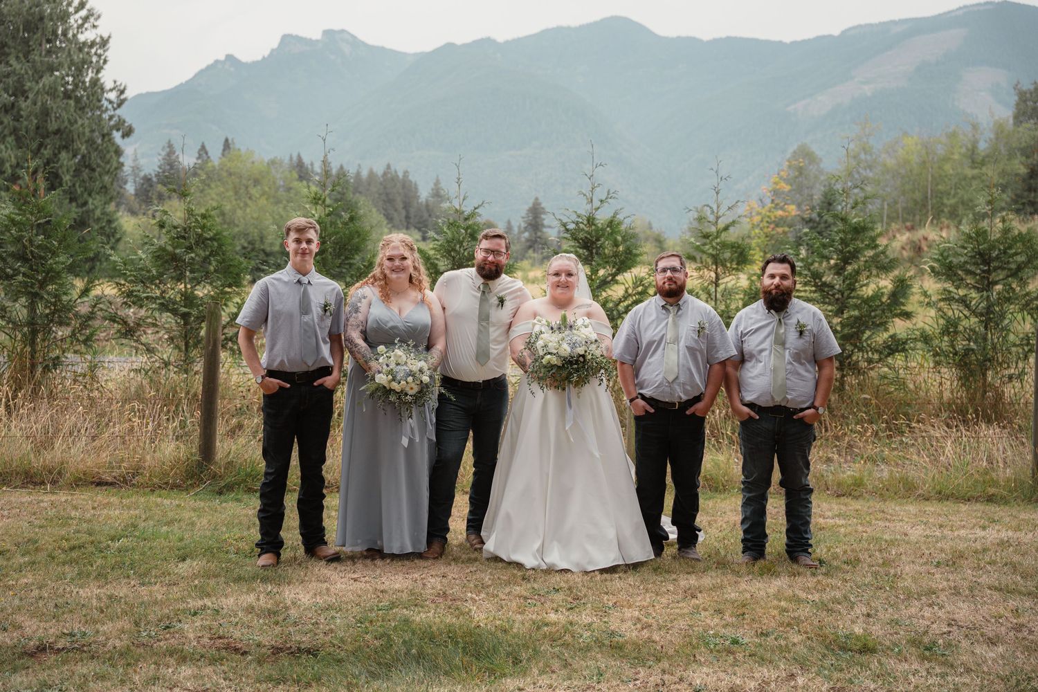 Wedding party poses outdoors with mountain and evergreen tree backdrop, bride in white gown flanked by groomsmen and bridesmaids.