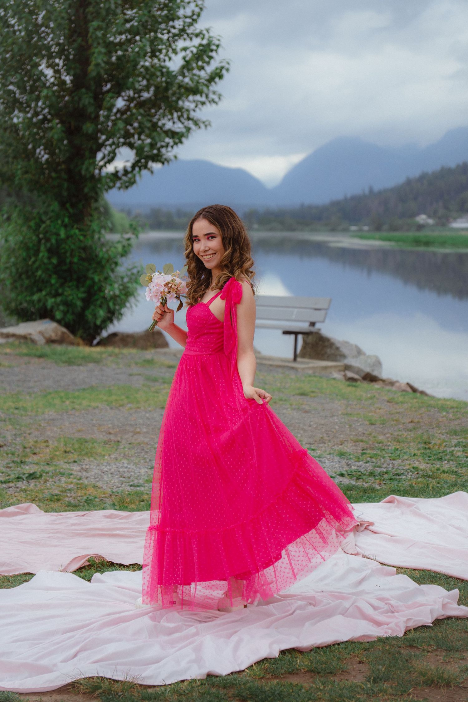 Smiling woman in hot pink formal dress holds bouquet beside reflective mountain lake.
