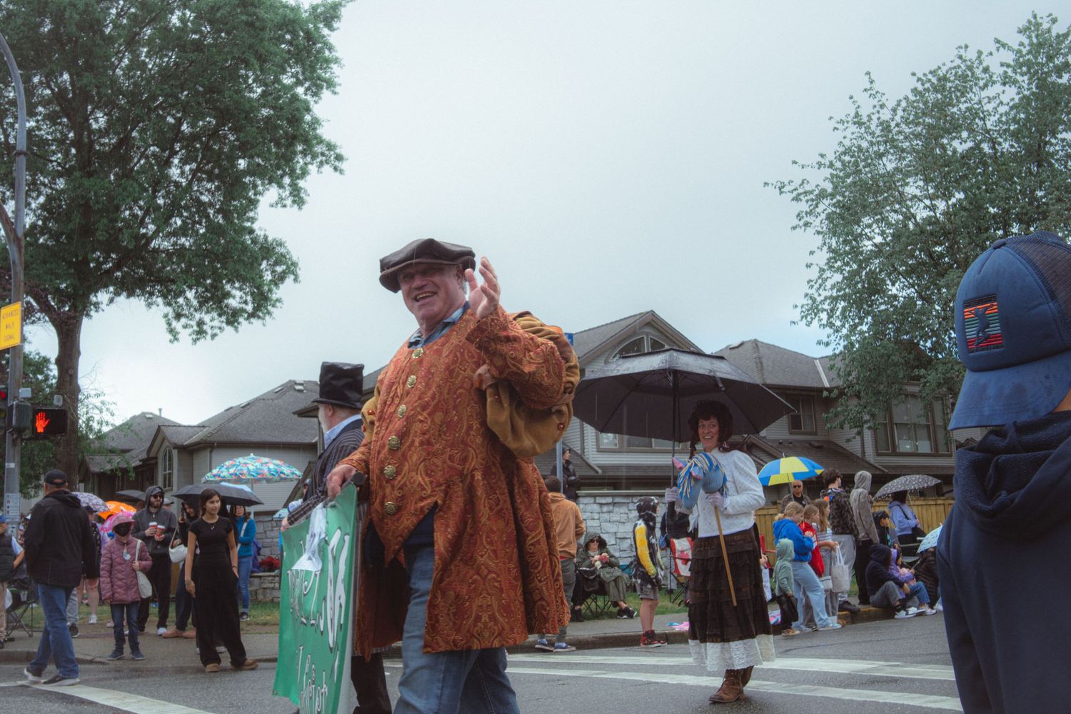 A man in a vintage brown coat walks through a lively street parade among costumed participants.