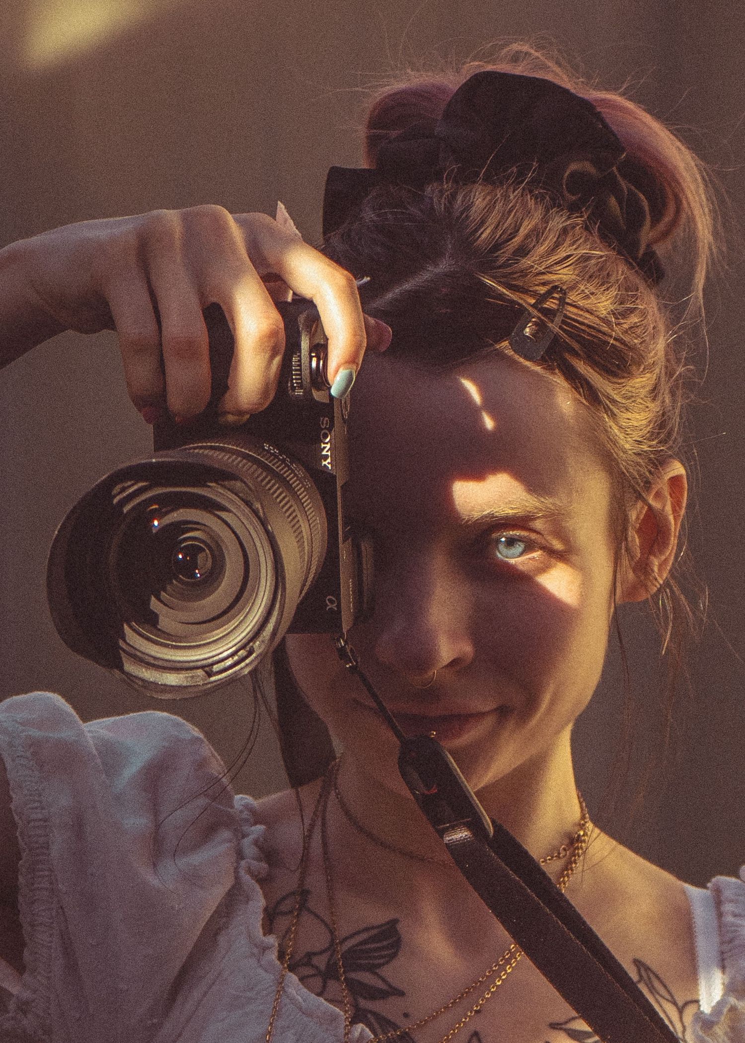 Young woman with tattoos holds DSLR camera up to face in warm sunlight with hair in bun.