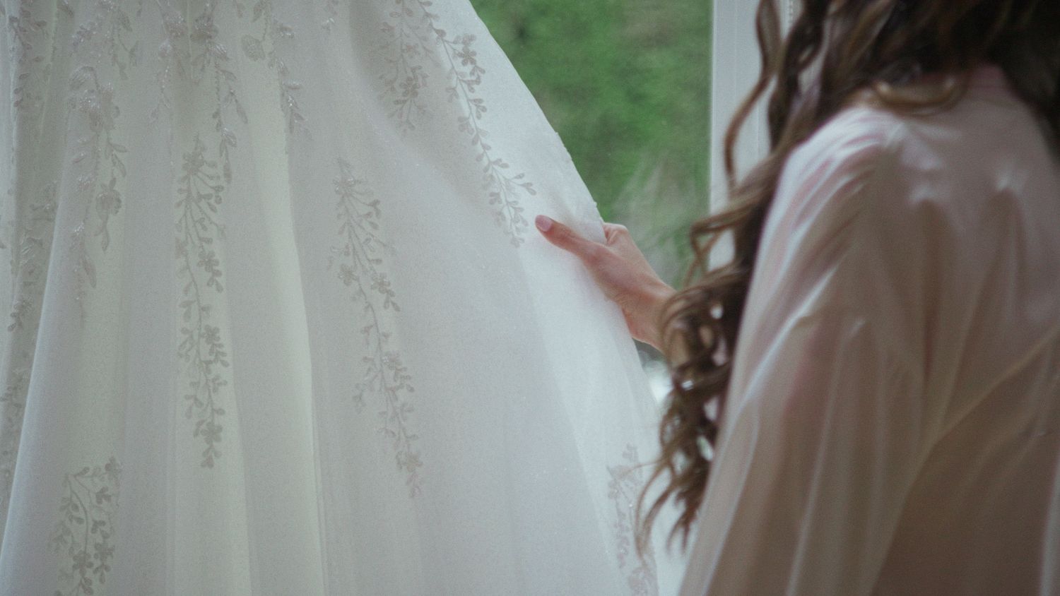 Bride in white robe gently touching elegant lace wedding dress hanging by window before ceremony.