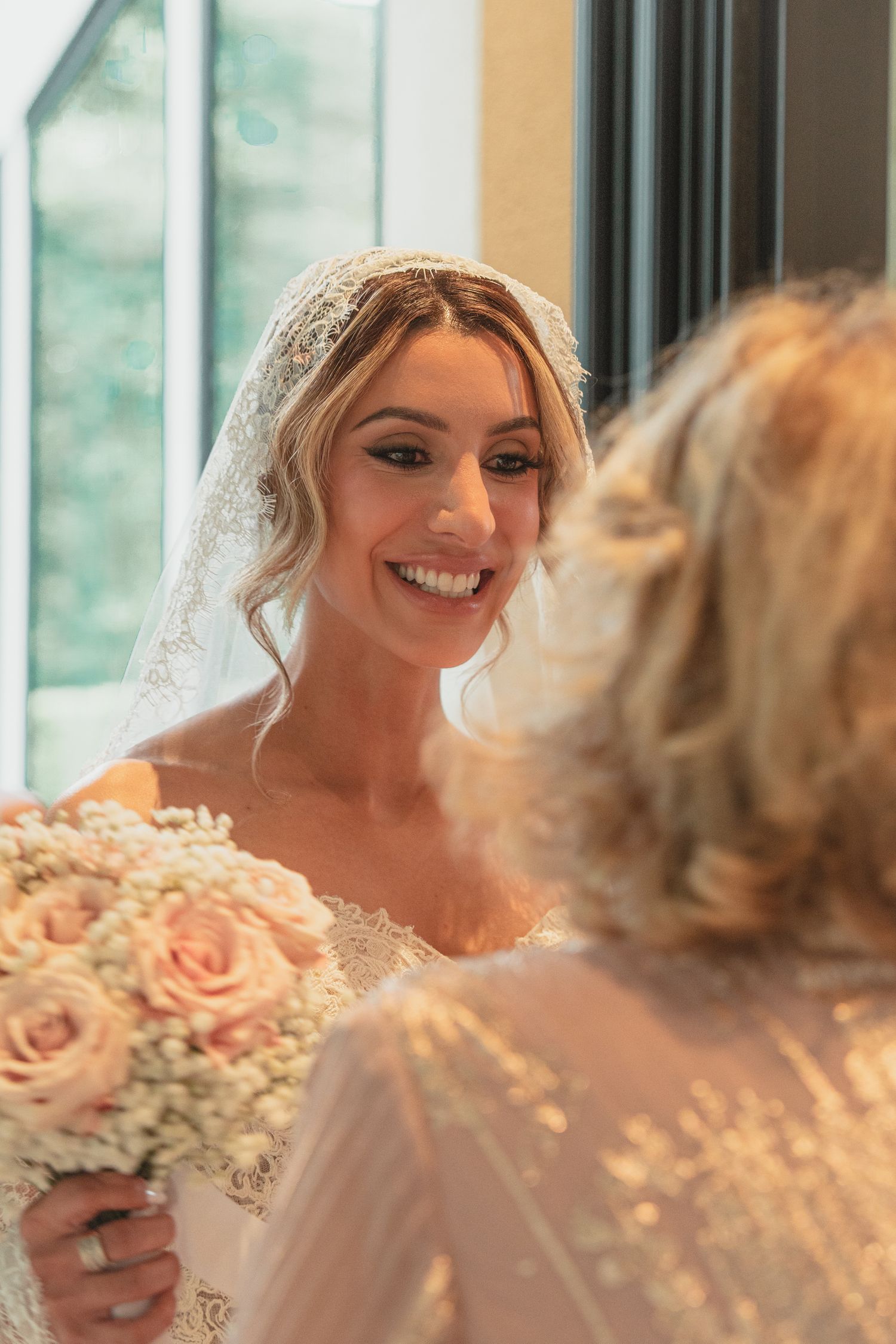 Smiling bride in lace veil holding blush pink rose bouquet on her wedding day.