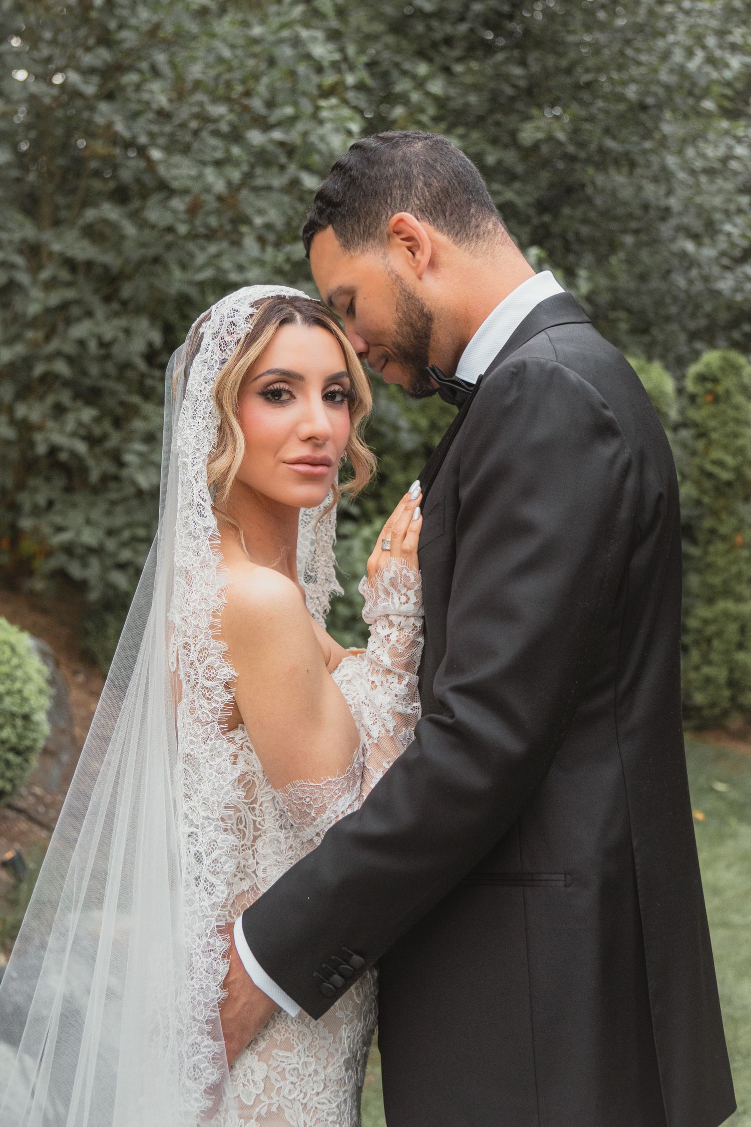 Bride in lace gown and veil embraces groom in black tuxedo outdoors among green trees.